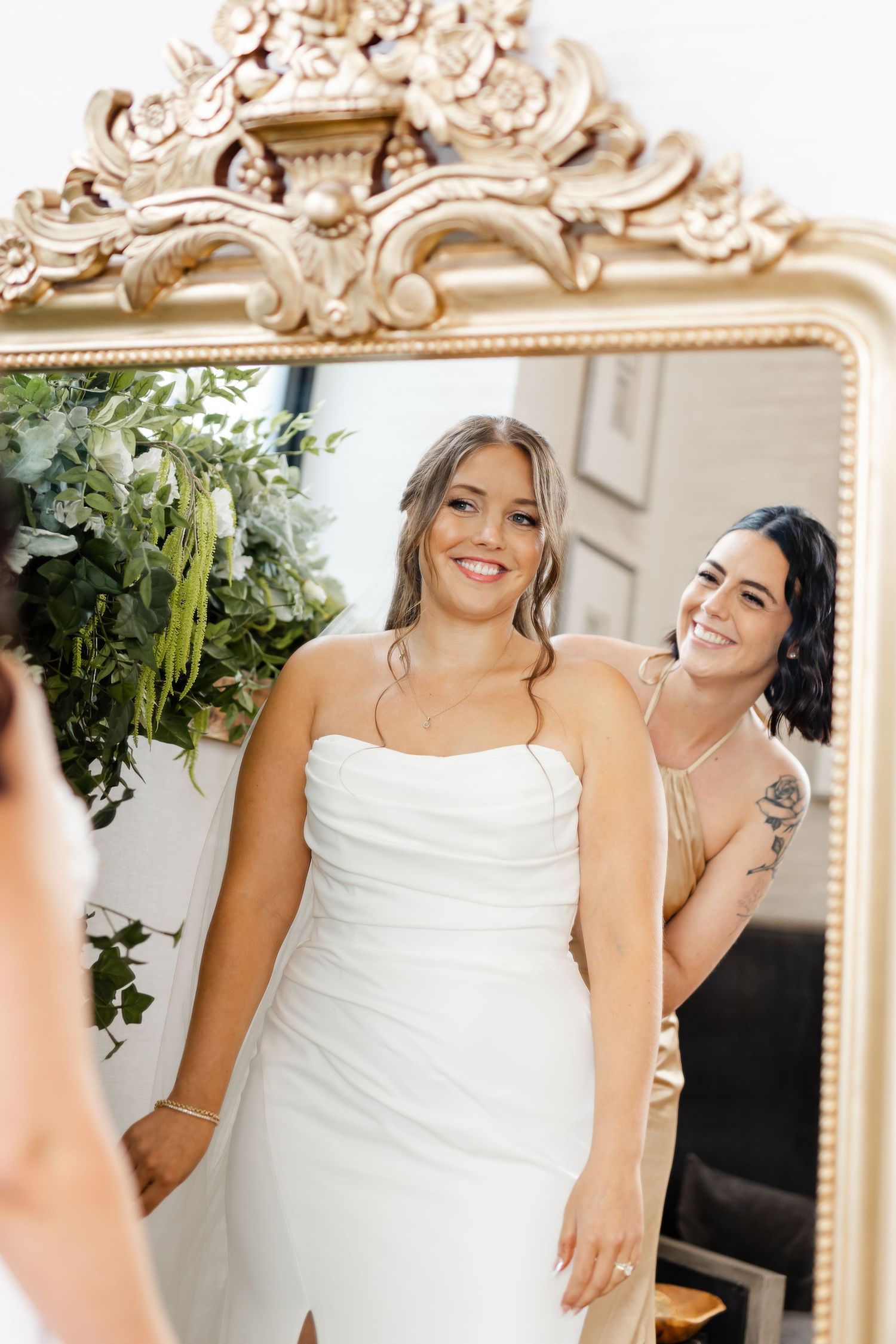 Bride smiles at ornate gold mirror reflection as bridesmaid with tattoo helps fasten her strapless white gown.