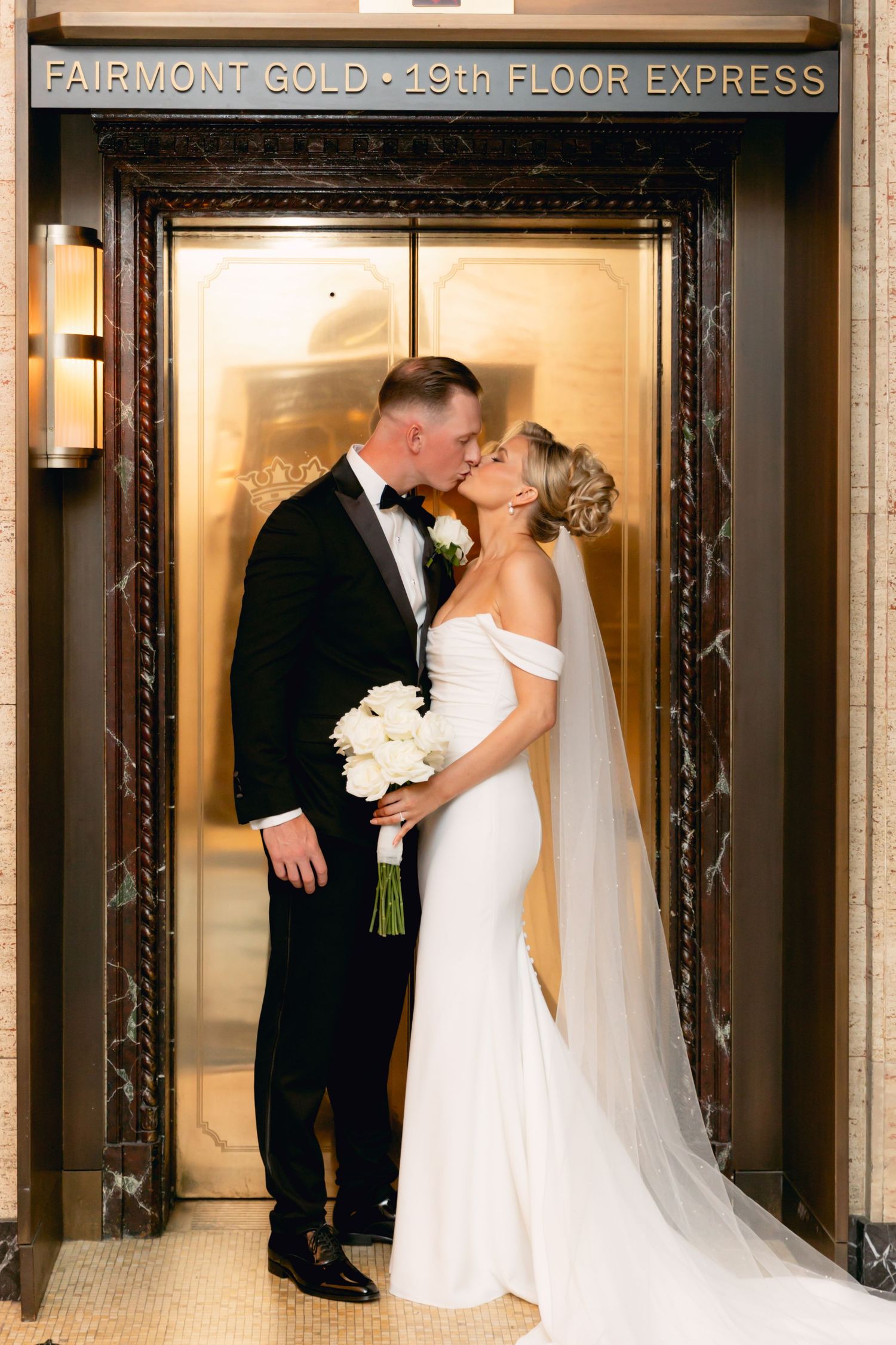 Bride and groom share a kiss at Fairmont Gold elevator, bride in fitted white gown holding white bouquet.