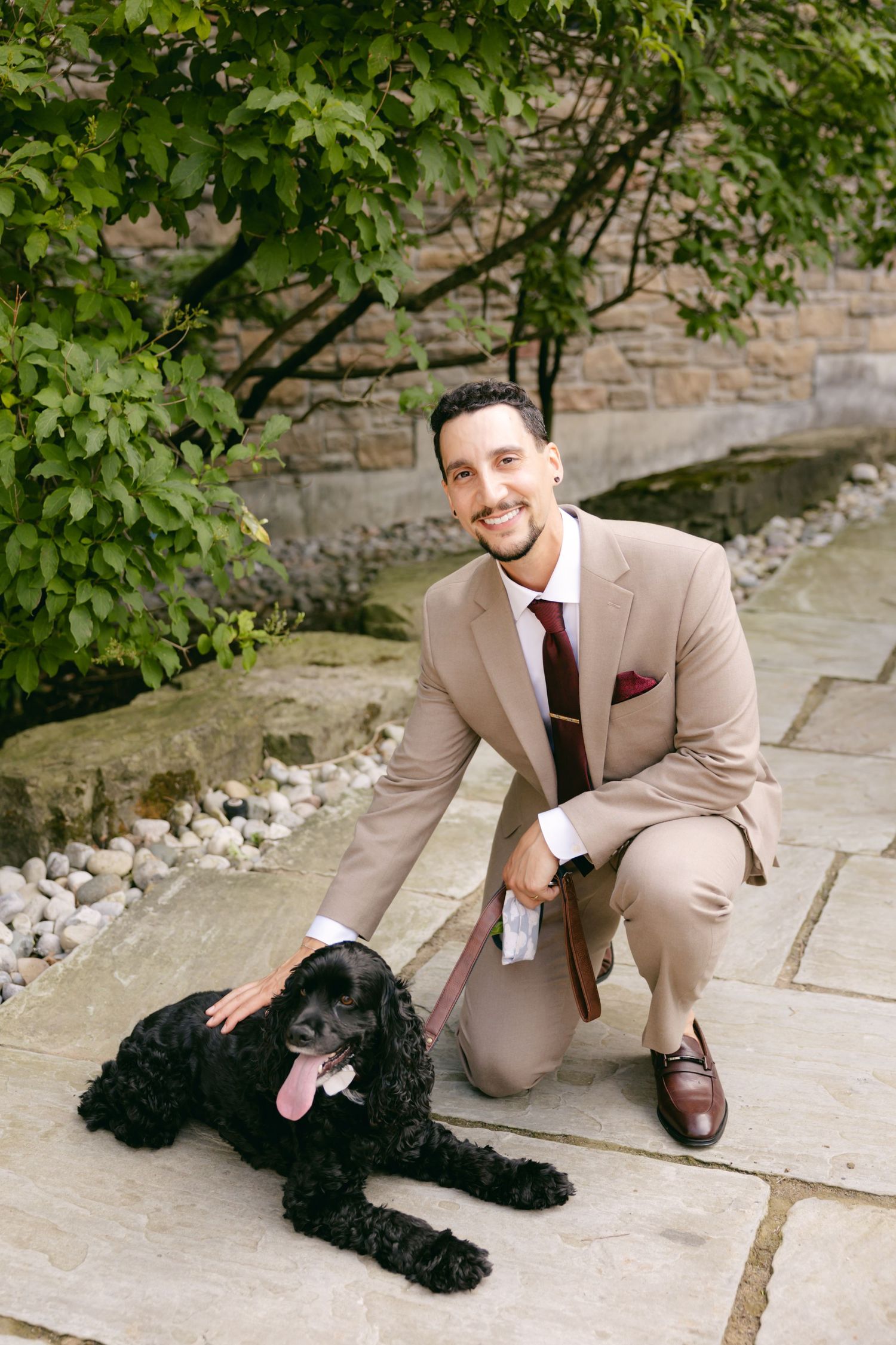 A groom in tan suit kneels beside black dog during outdoor wedding photos.