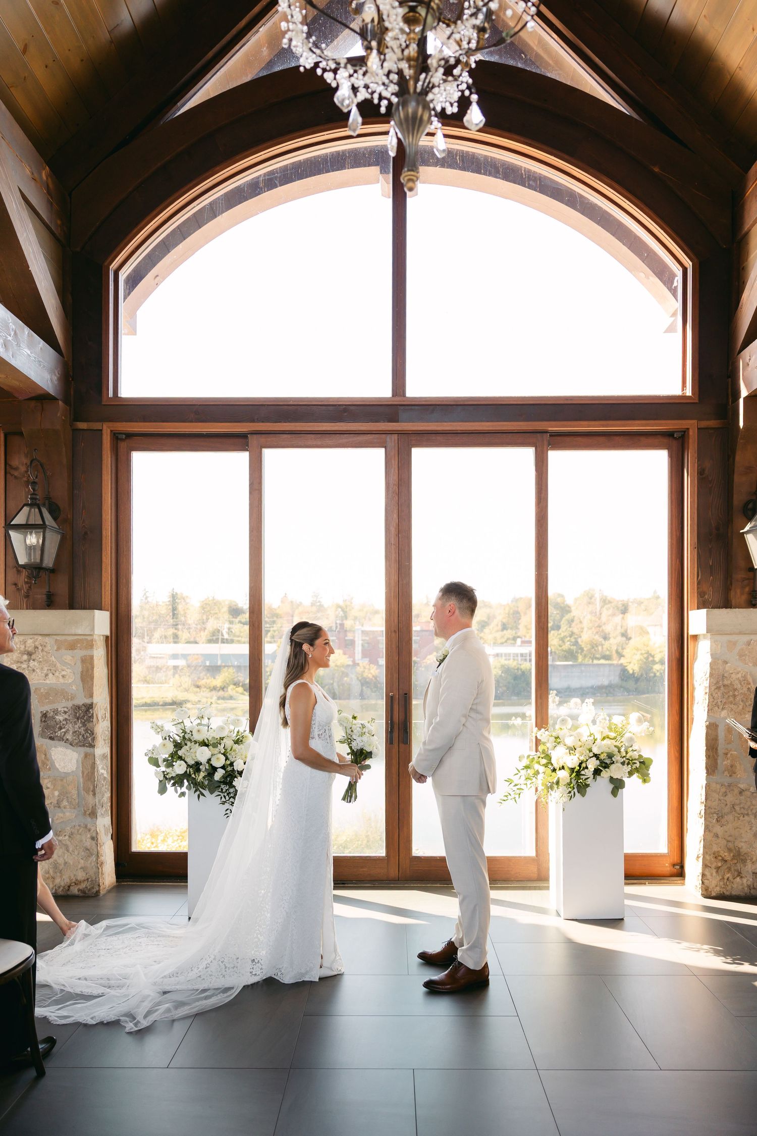 A bride and groom stand before large windows in an elegant venue with vaulted ceilings and stone accents.