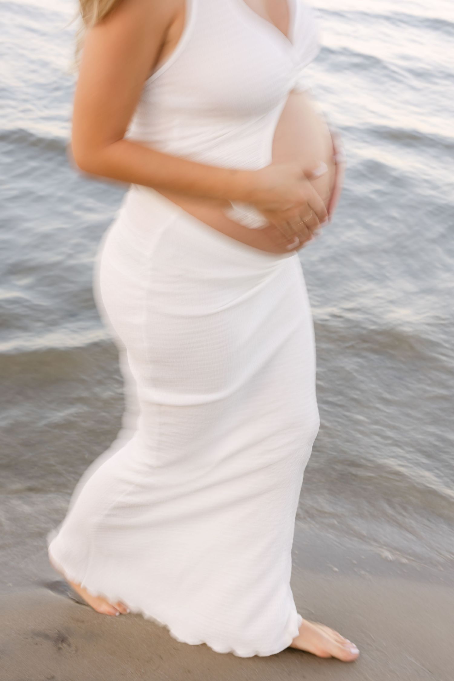 Woman in flowing white dress walking along beach shoreline at sunset.