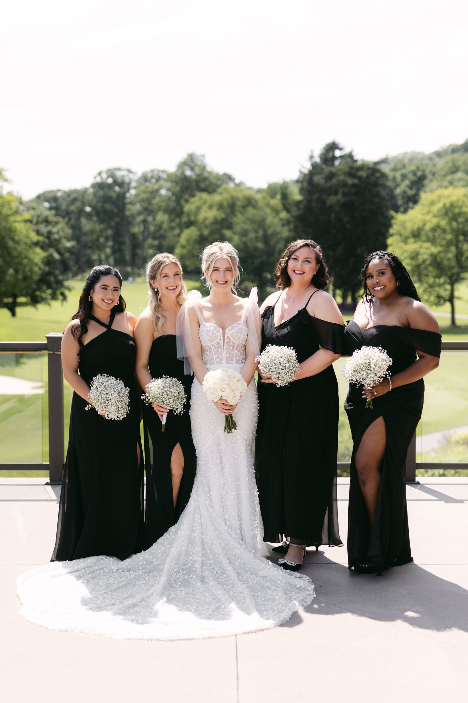 Wedding party in black dresses poses with bride in white lace gown holding white bouquets on sunny patio.