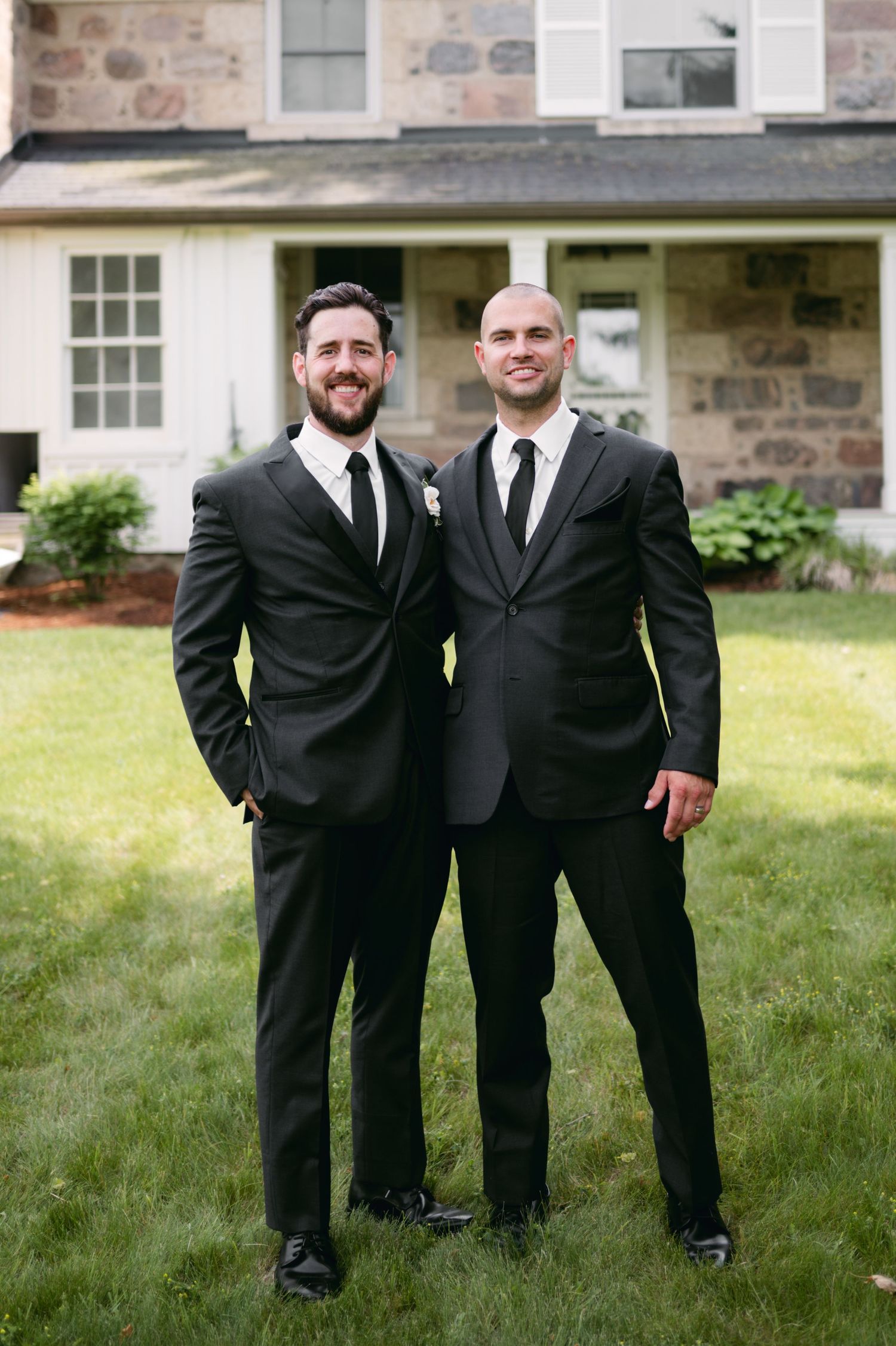 Two wedding party members in black suits stand together on lawn near stone building.