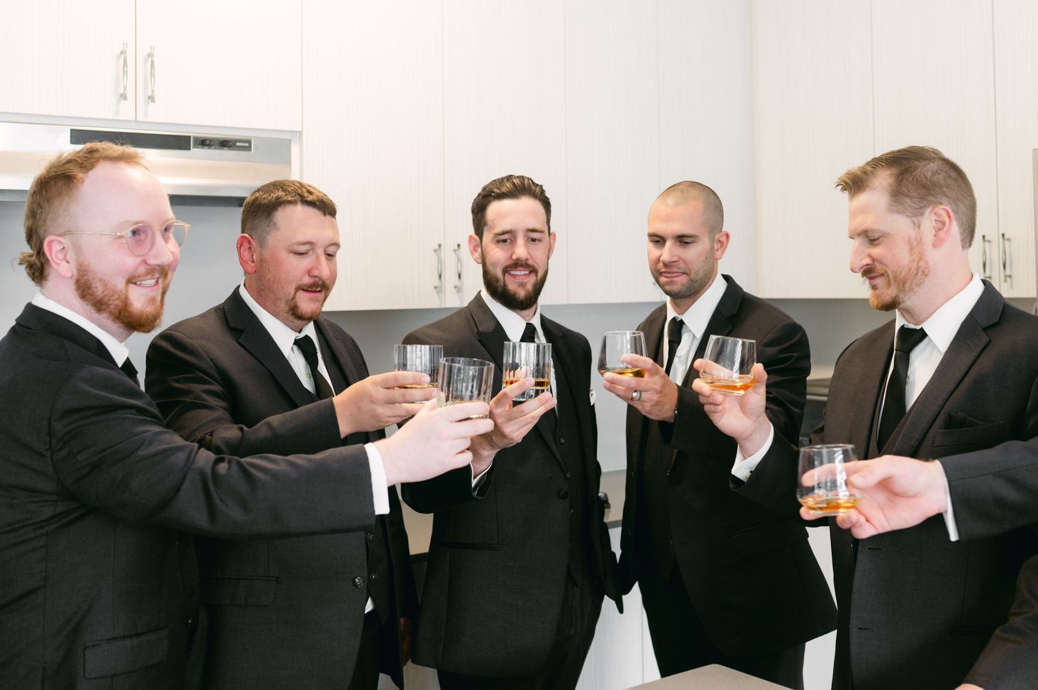 Groomsmen in black suits share drinks during pre-wedding celebrations.
