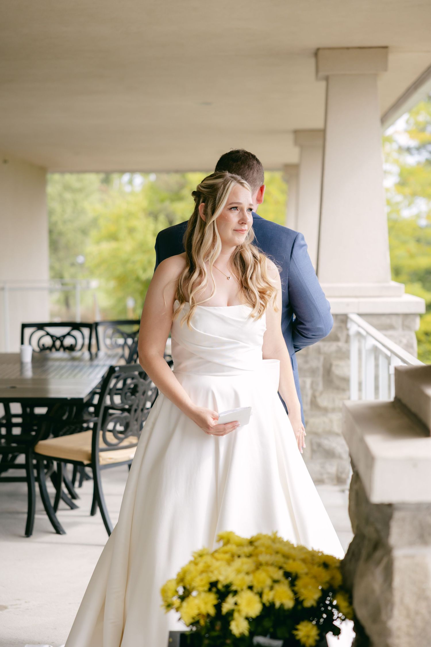 Bride in elegant white gown stands on covered porch with yellow flowers in foreground.