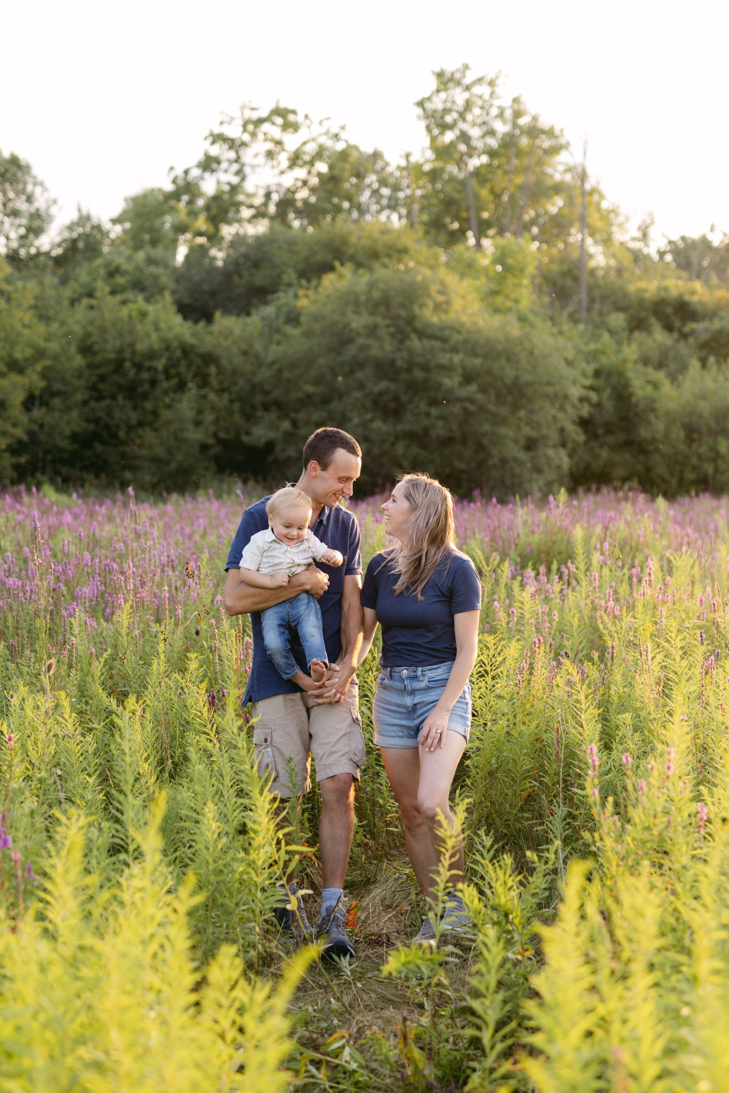 A young family stands in a vibrant purple wildflower field, with the father holding a baby while gazing lovingly at the mother.