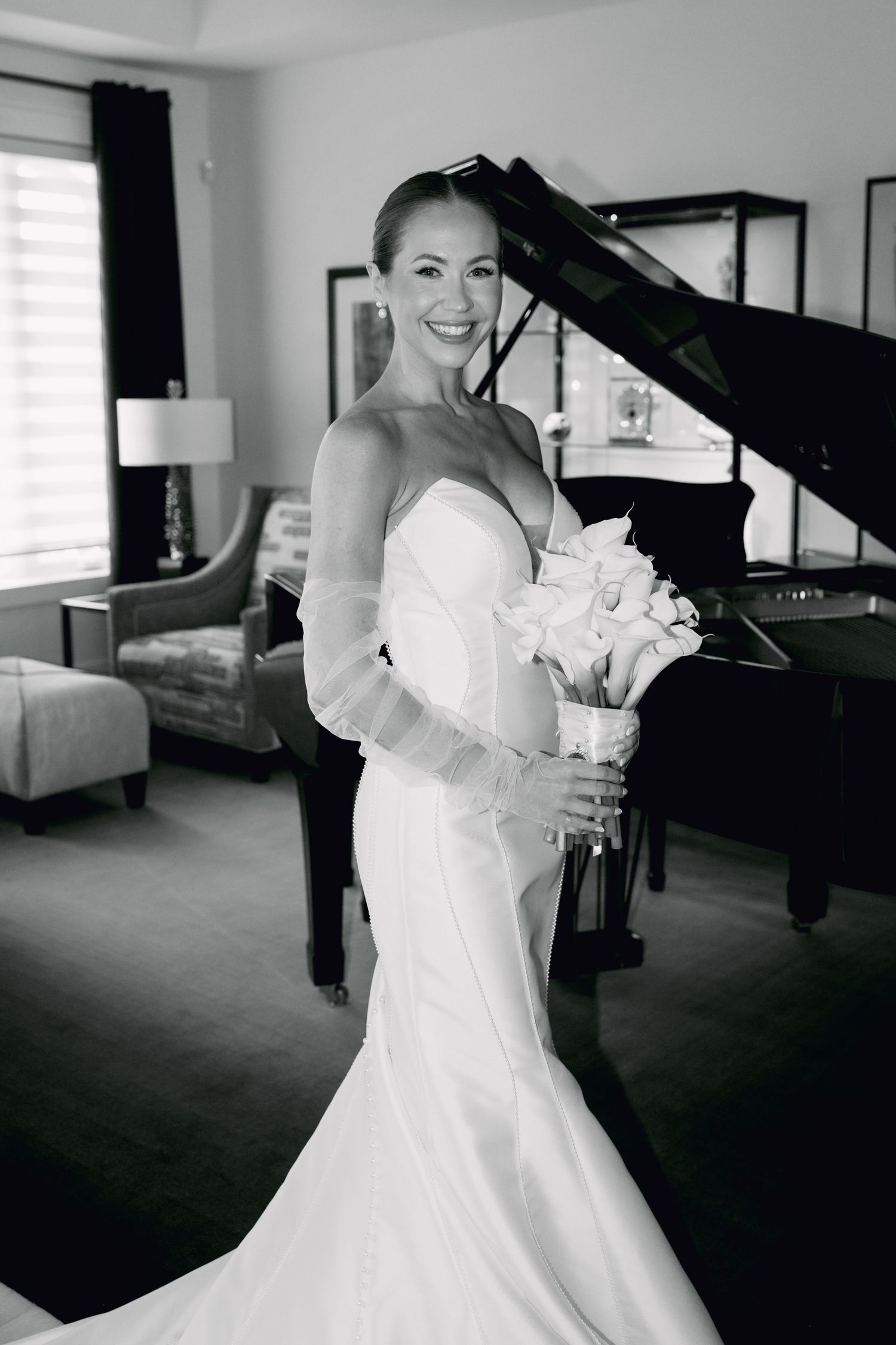 Smiling bride in strapless white gown holding bouquet stands before grand piano in elegant room, black and white photo.