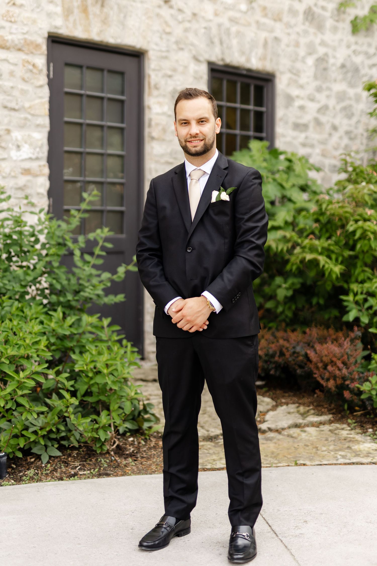 Groom in black suit with white boutonniere stands confidently outside stone building surrounded by lush greenery.