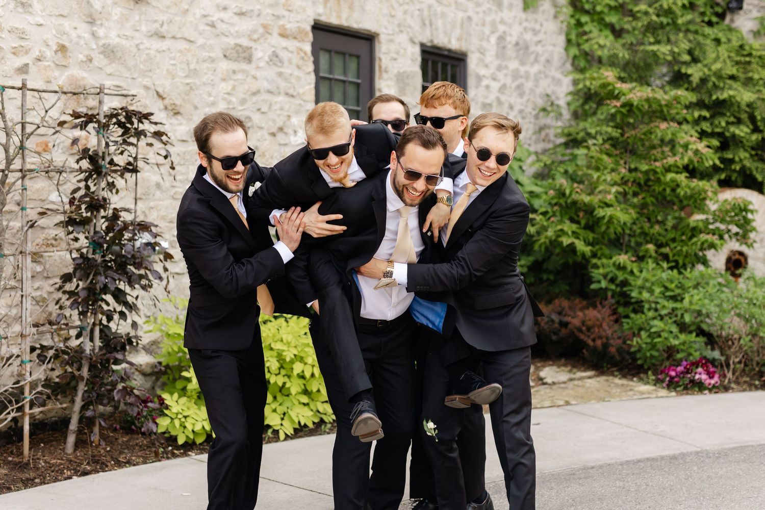 Groomsmen in black suits and sunglasses playfully lift groom outdoors near stone building on wedding day.