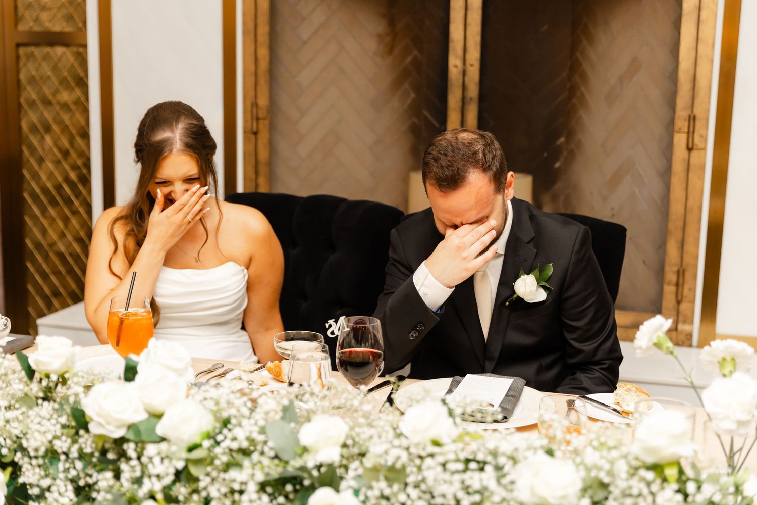 Bride and groom cover their faces laughing emotionally during heartfelt wedding reception speeches at head table.