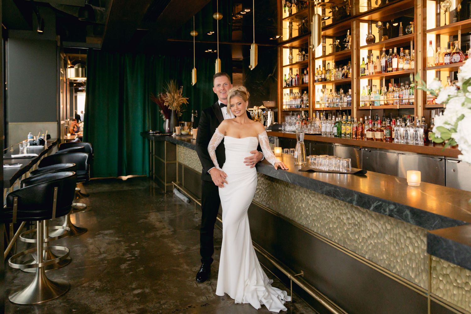 Bride and groom pose together at a sophisticated bar with green velvet walls and gold accents.