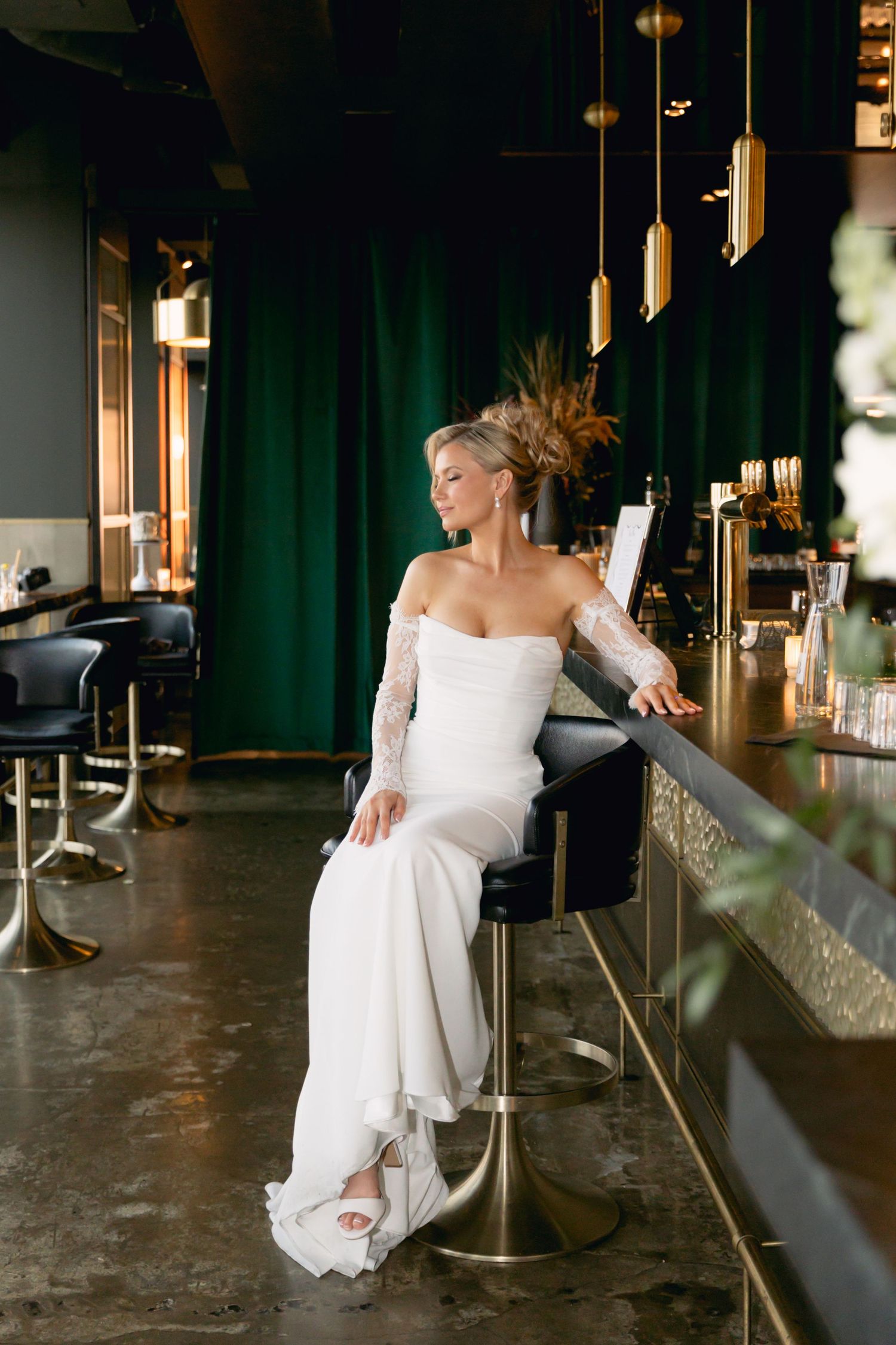 Elegant bride in white lace gown seated at a dark bar with green curtains and gold pendant lights.