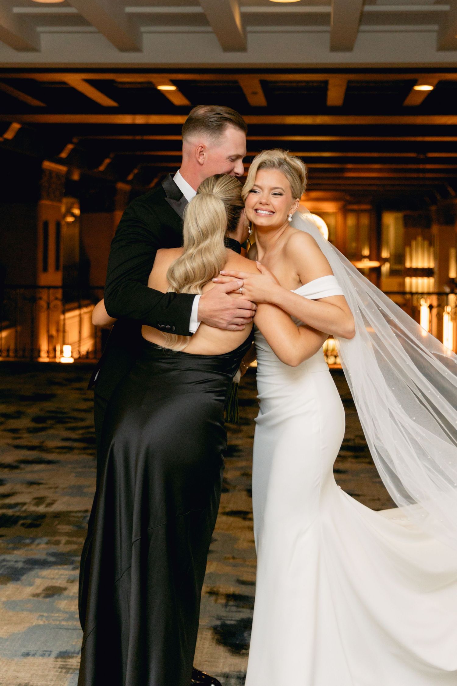 Bride in white gown and veil shares a group hug with groom in tuxedo and guest in black dress at elegant wedding reception.