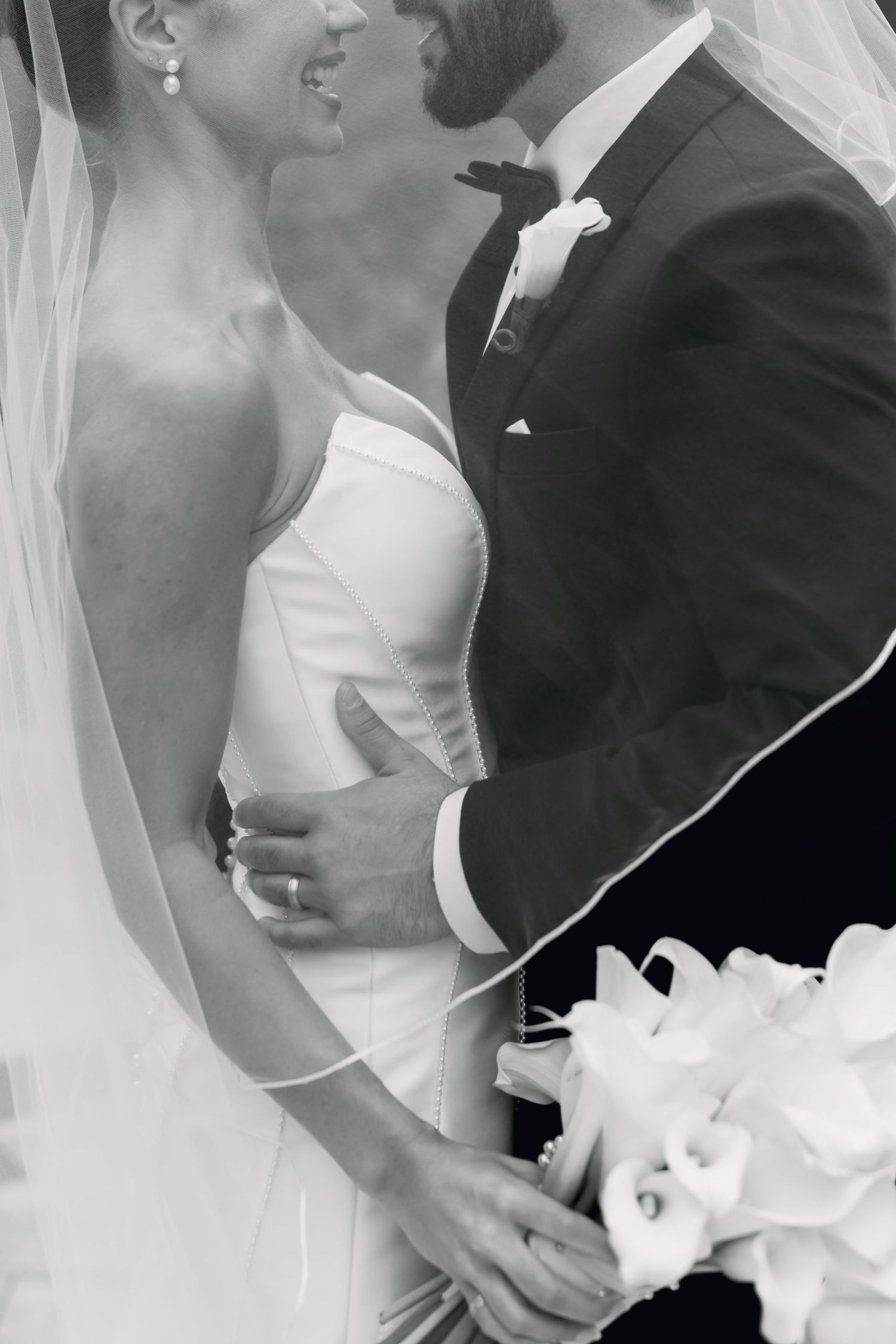 Black and white close-up of bride and groom embracing, bride holding white calla lily bouquet with veil draped.