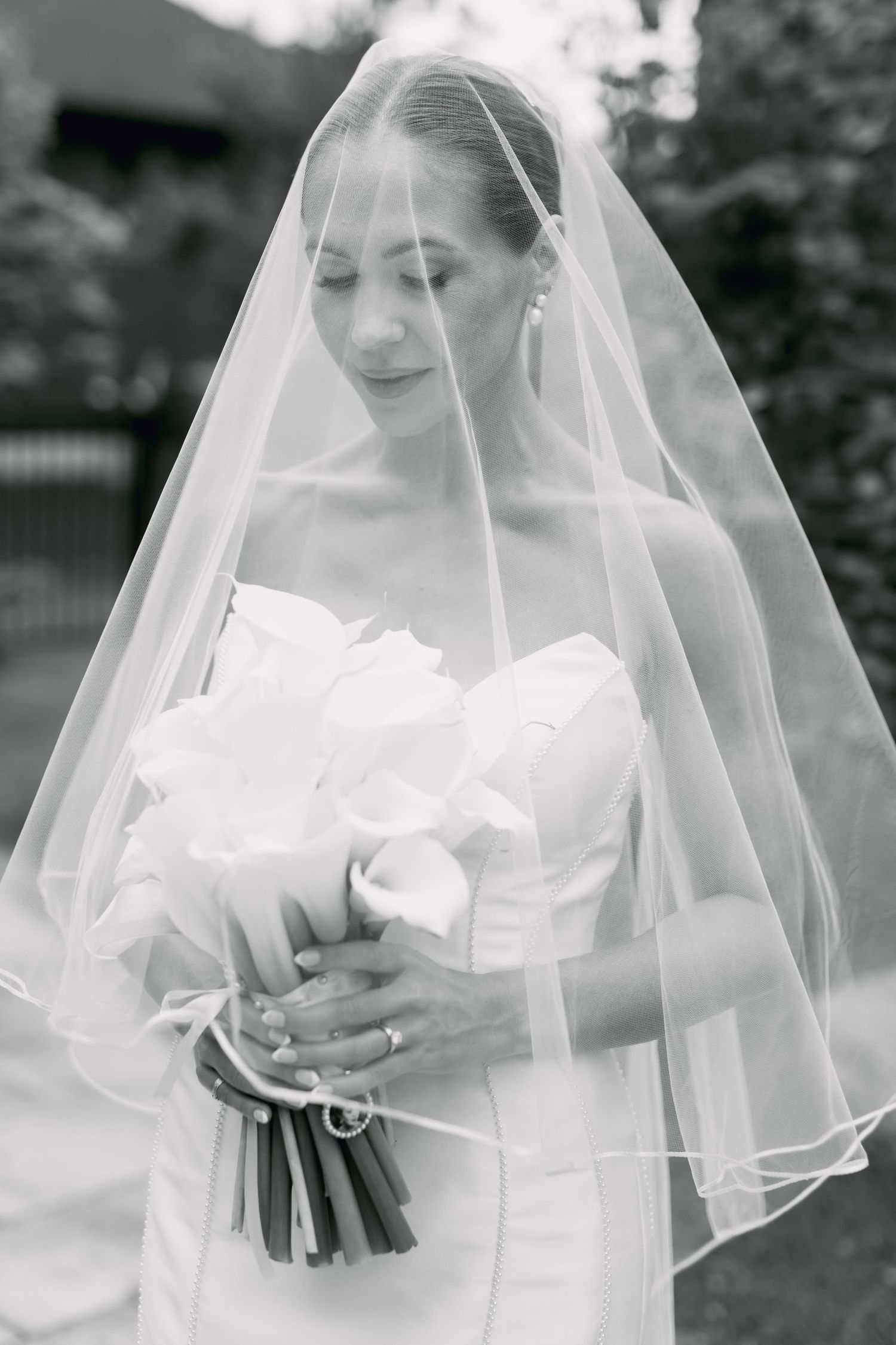 Black and white portrait of bride in veil holding white calla lily bouquet, eyes downcast.