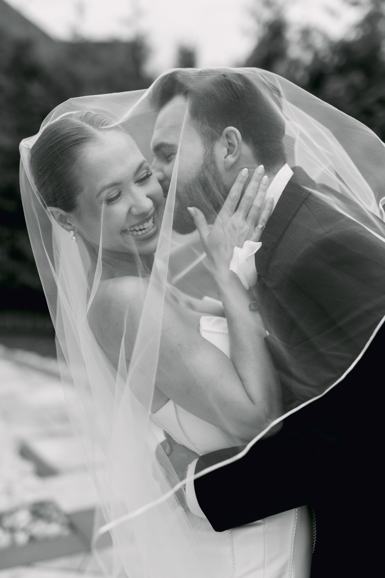 Black and white photo of a smiling bride and groom sharing a romantic moment under a wedding veil.