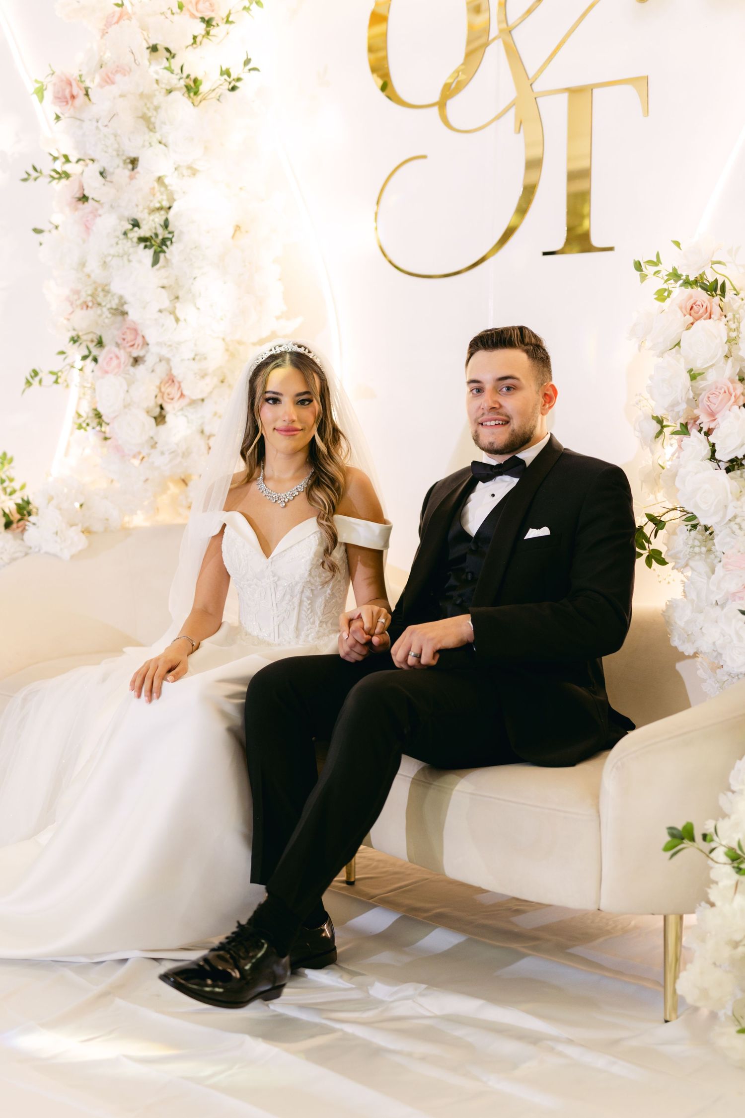 Newlyweds pose together on white furniture against a floral backdrop with gold lettering.