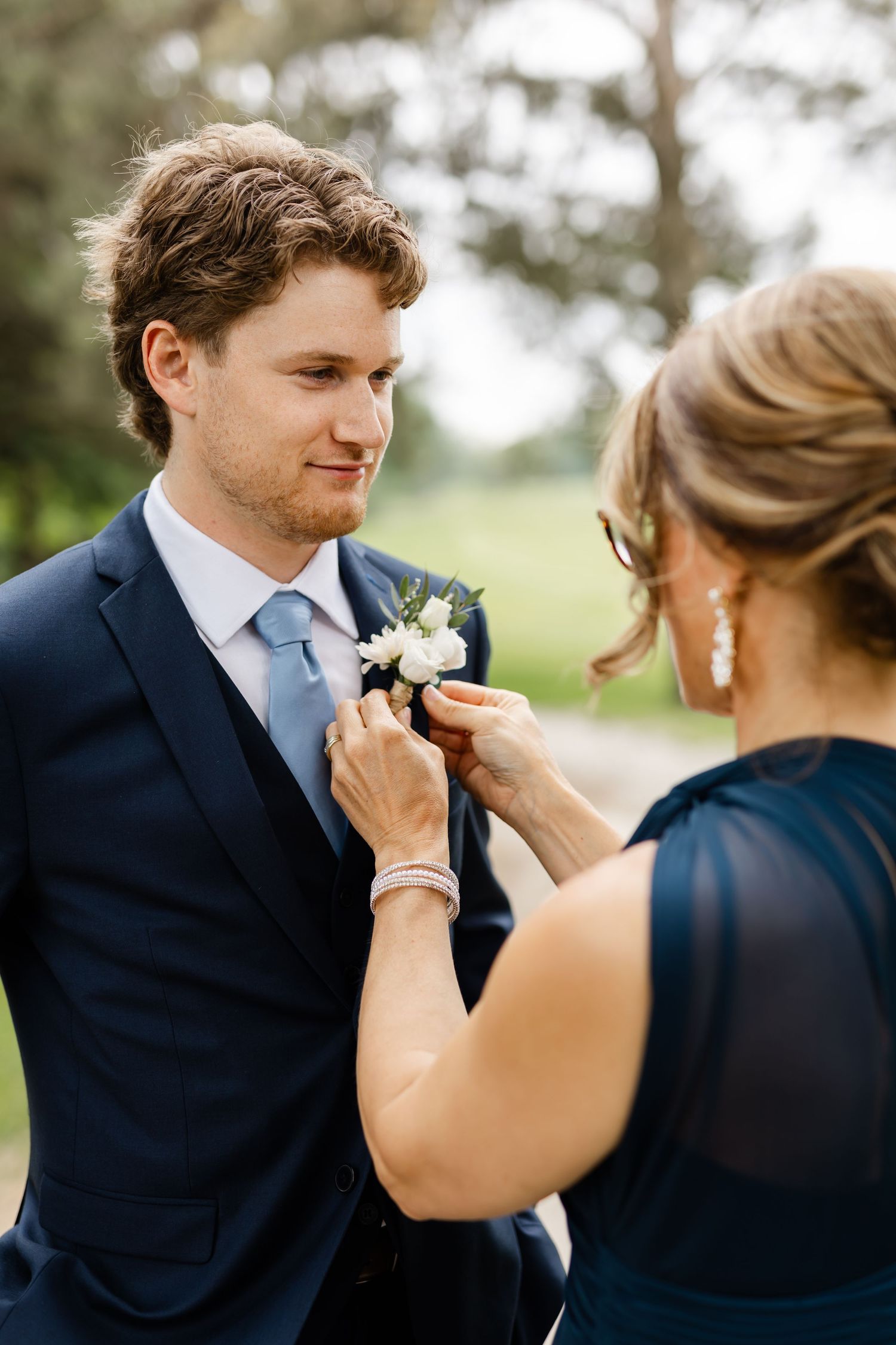 Wedding party member helps adjust a navy blue suit and boutonniere in an outdoor setting.