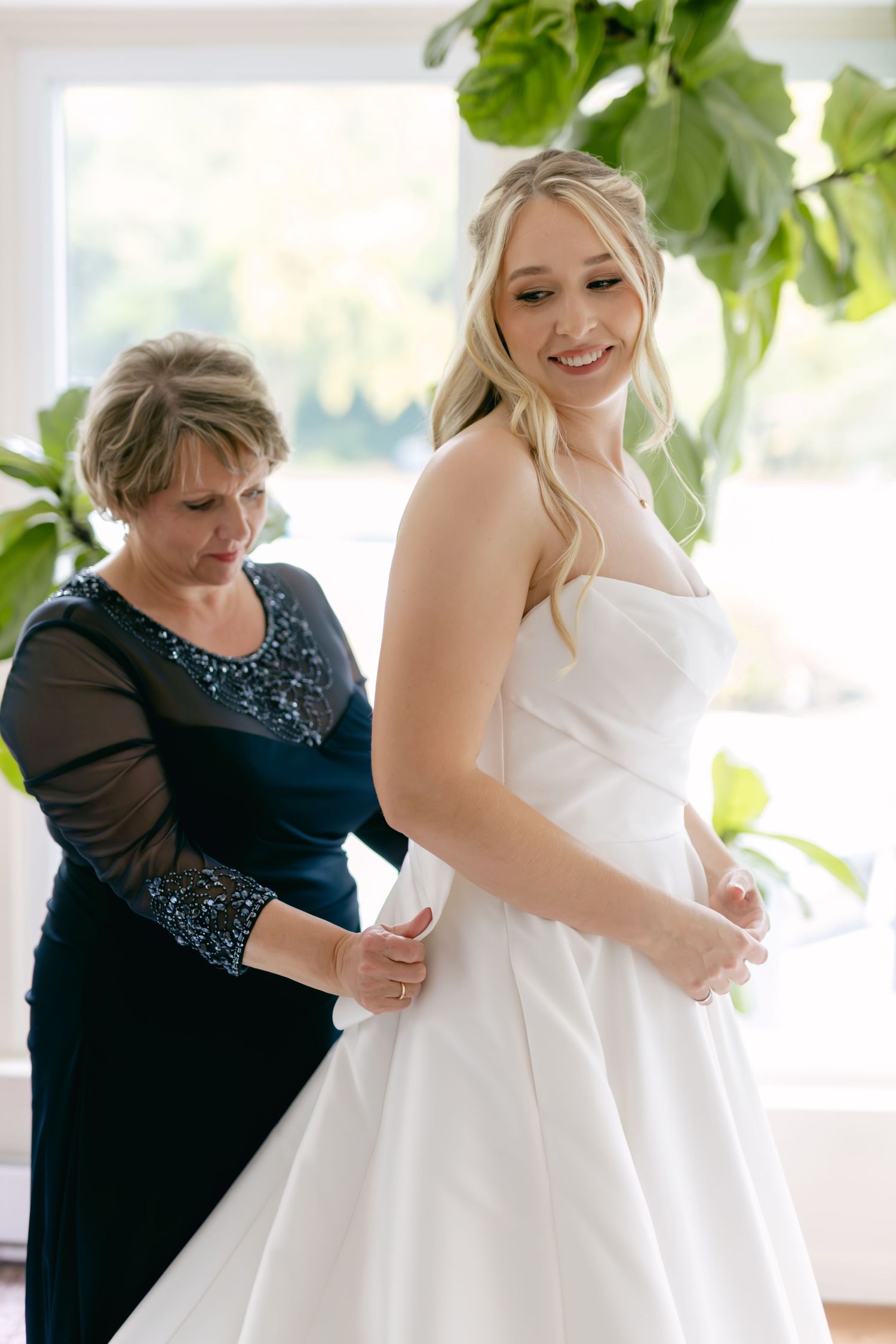 Helper adjusts the flowing train of an elegant white wedding gown in a sunlit room with green plants.
