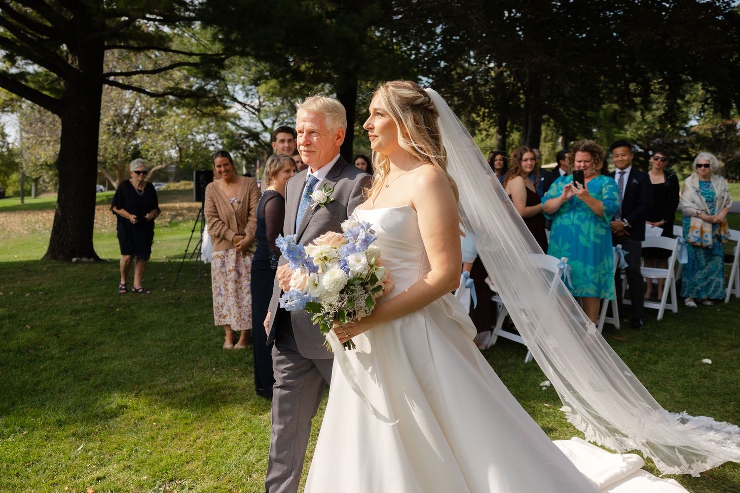Wedding processional on grassy lawn with white floral decorations and seated guests.