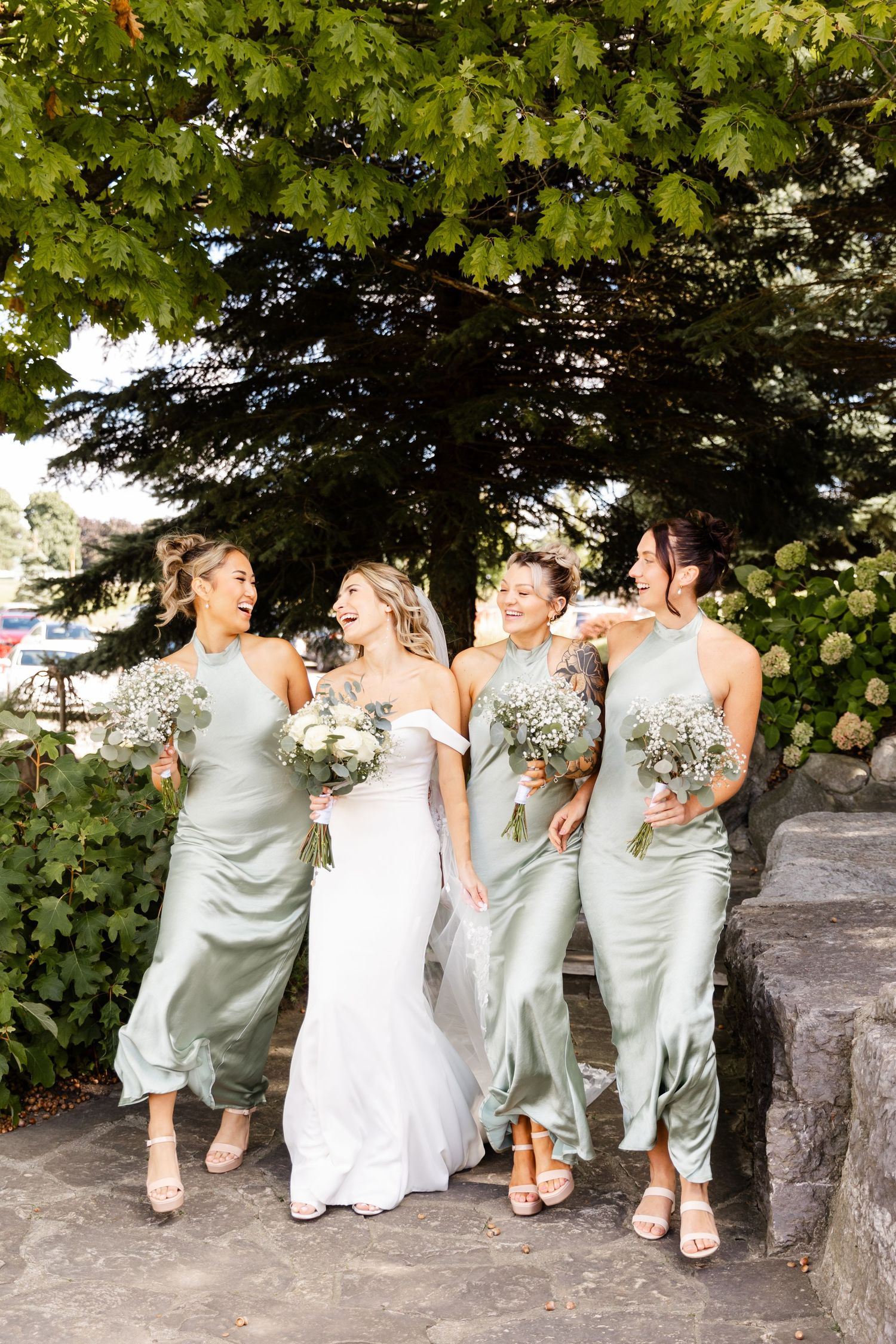 Wedding party in sage green dresses and white gown pose playfully on stone wall under green foliage.