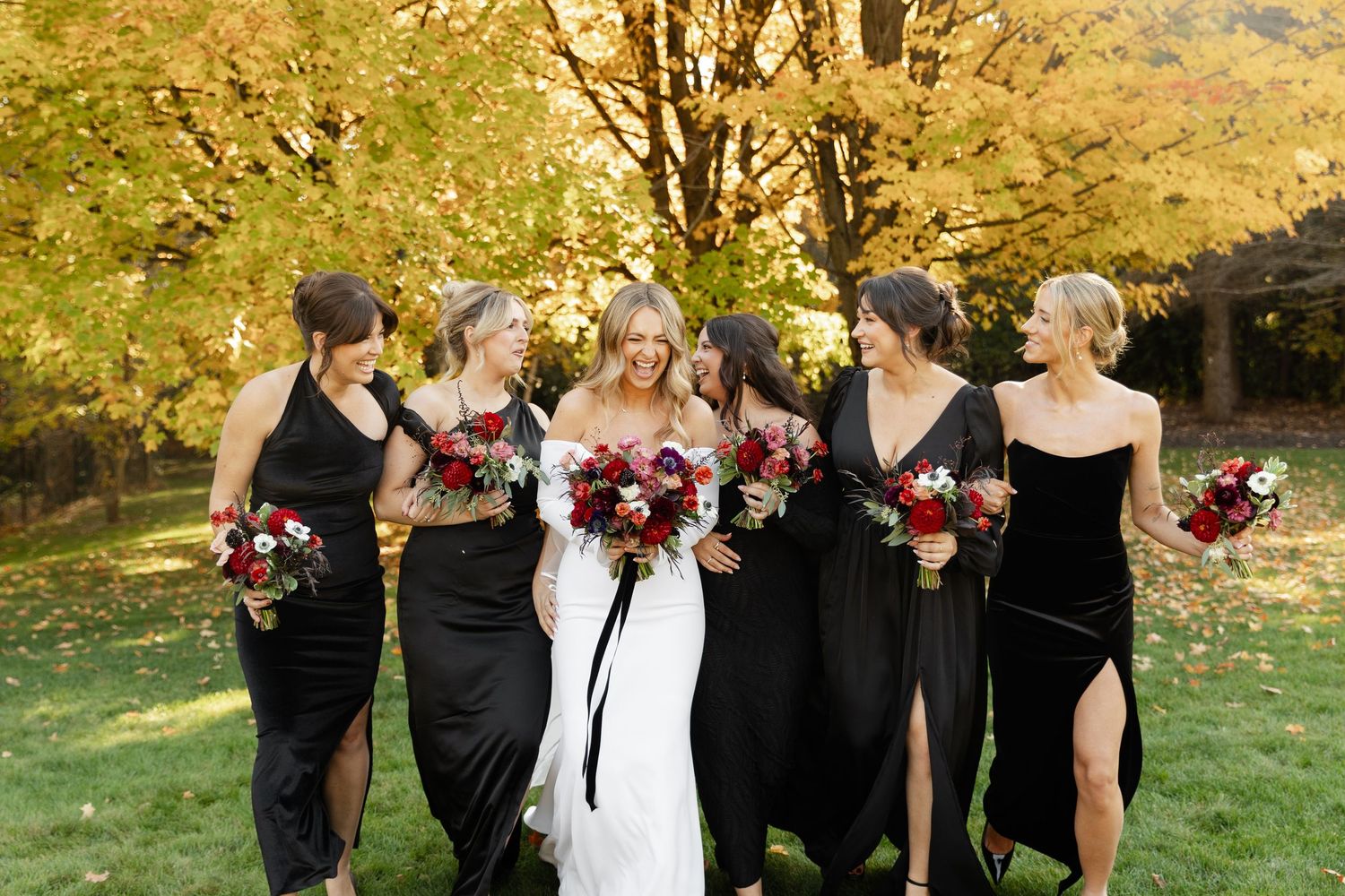 Wedding party in black dresses poses outdoors against autumn foliage.