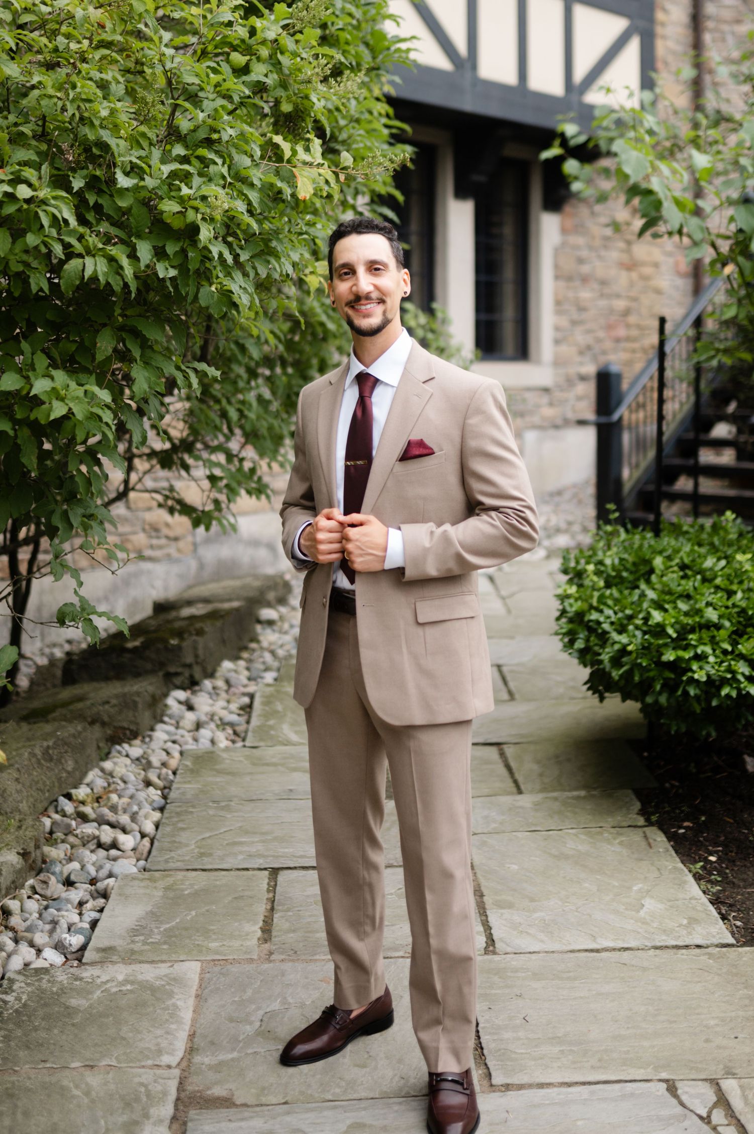 Well-dressed person in beige suit stands confidently on stone pathway near landscaped garden.