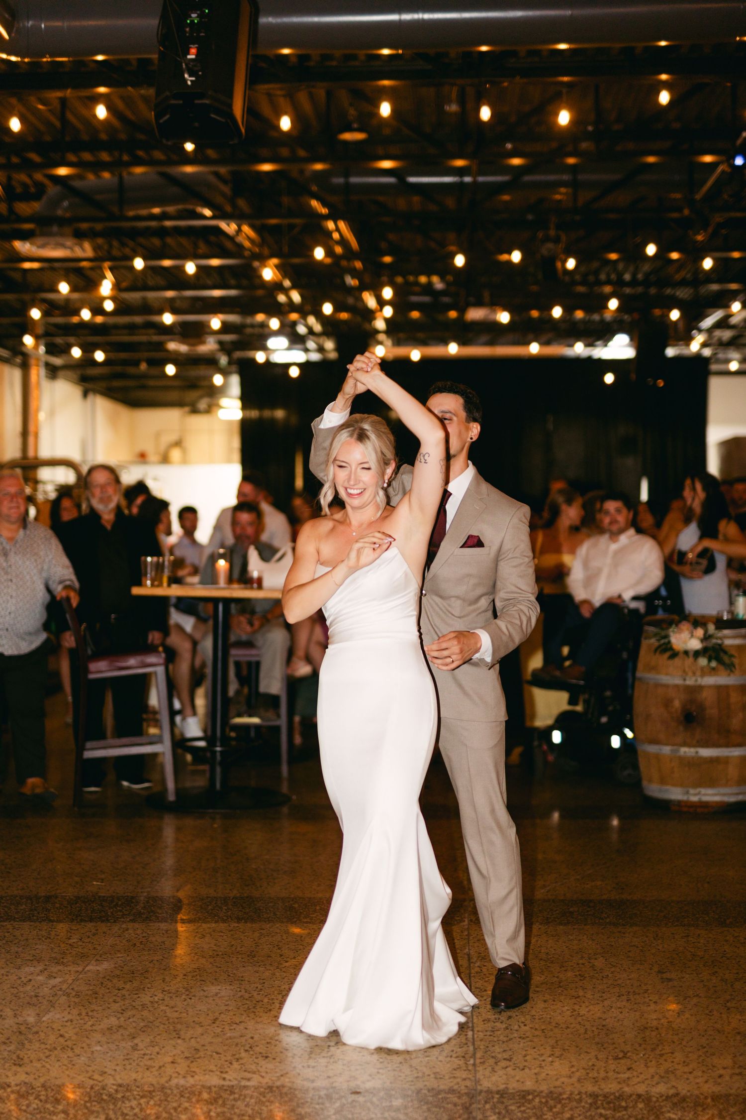 Bride dancing with guests during wedding reception under industrial string lights.