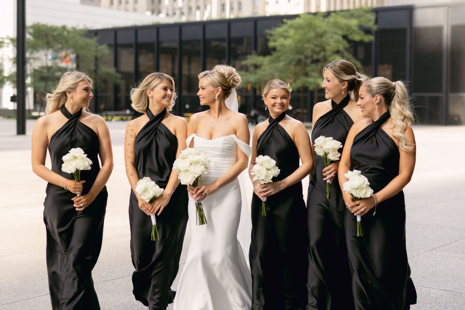 Bride in white gown with five bridesmaids in black halter dresses holding white bouquets outdoors.