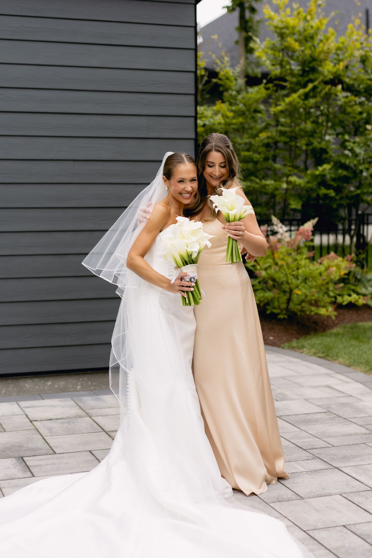 Smiling bride in white strapless gown and veil poses with bridesmaid in champagne dress holding bouquets.