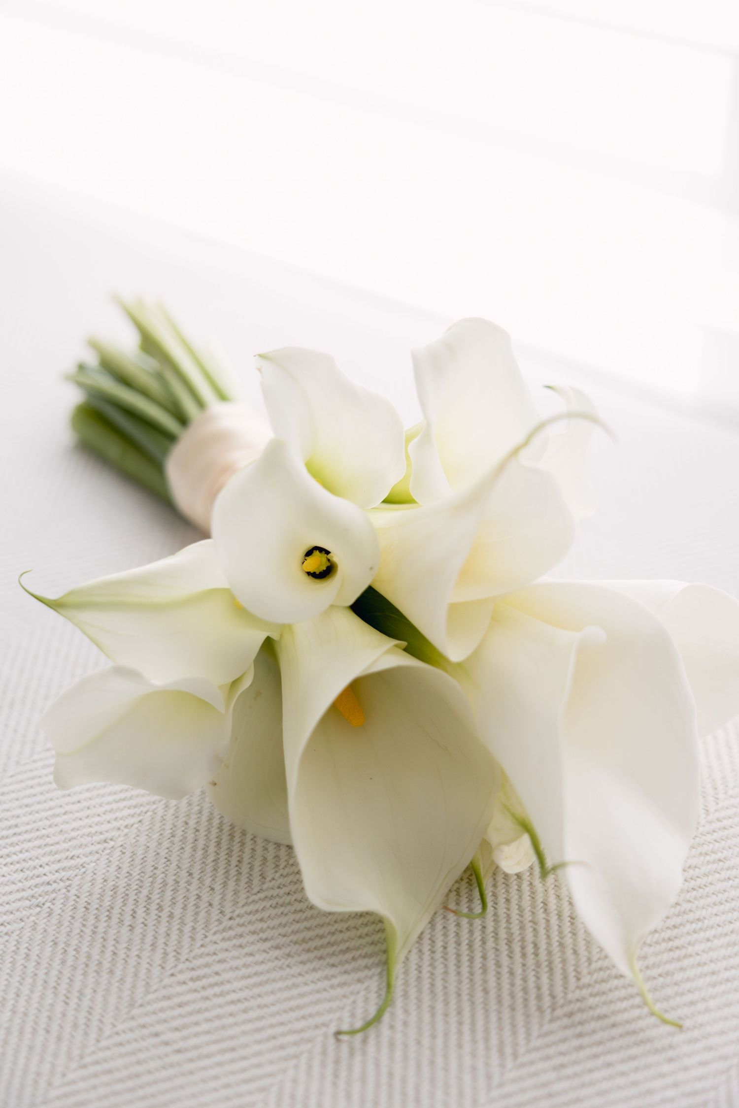 Elegant white calla lily bridal bouquet tied with ribbon resting on white textured surface.