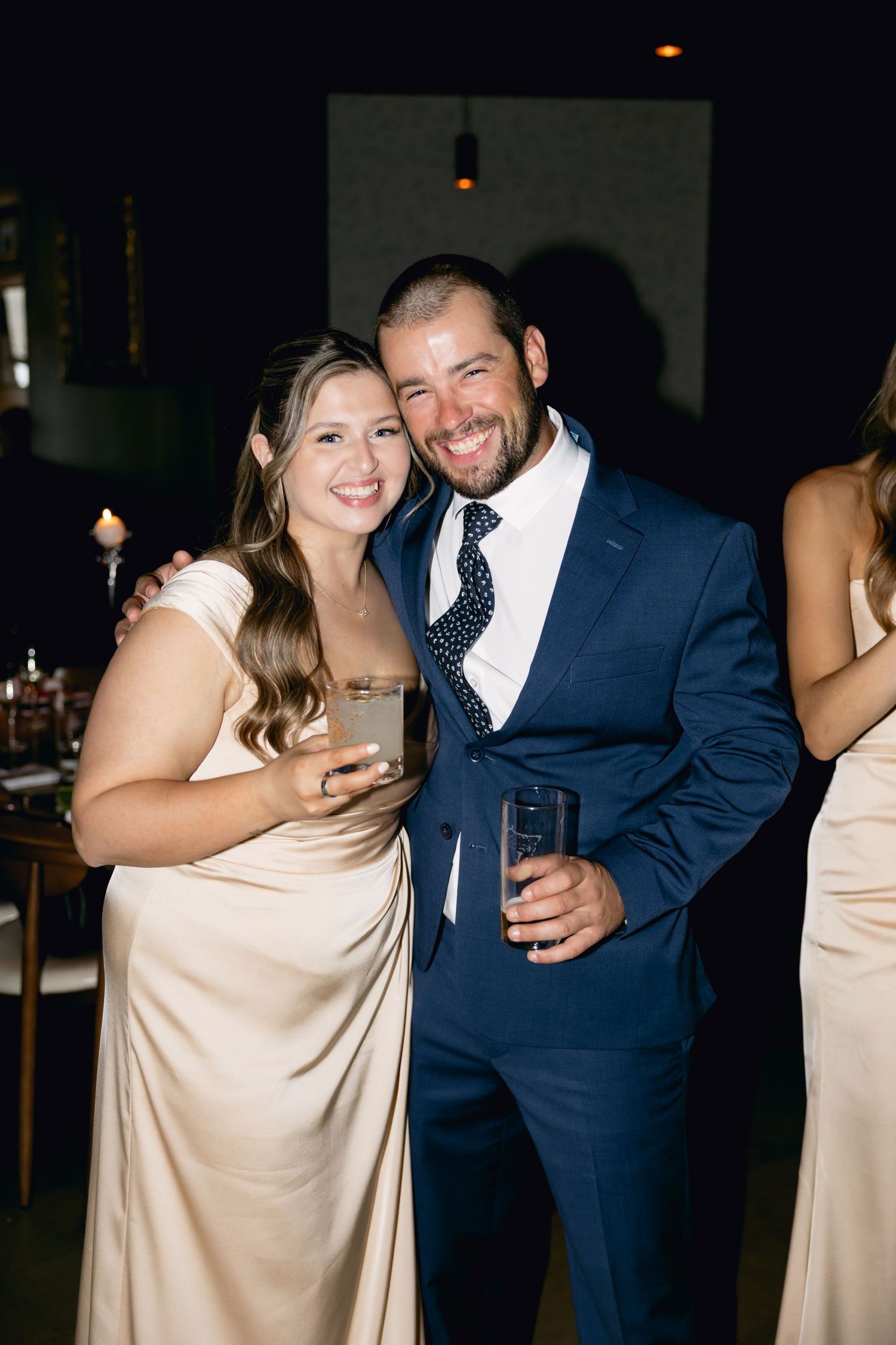 Smiling couple at wedding reception, woman in champagne gown holding drink, man in navy suit with polka dot tie.