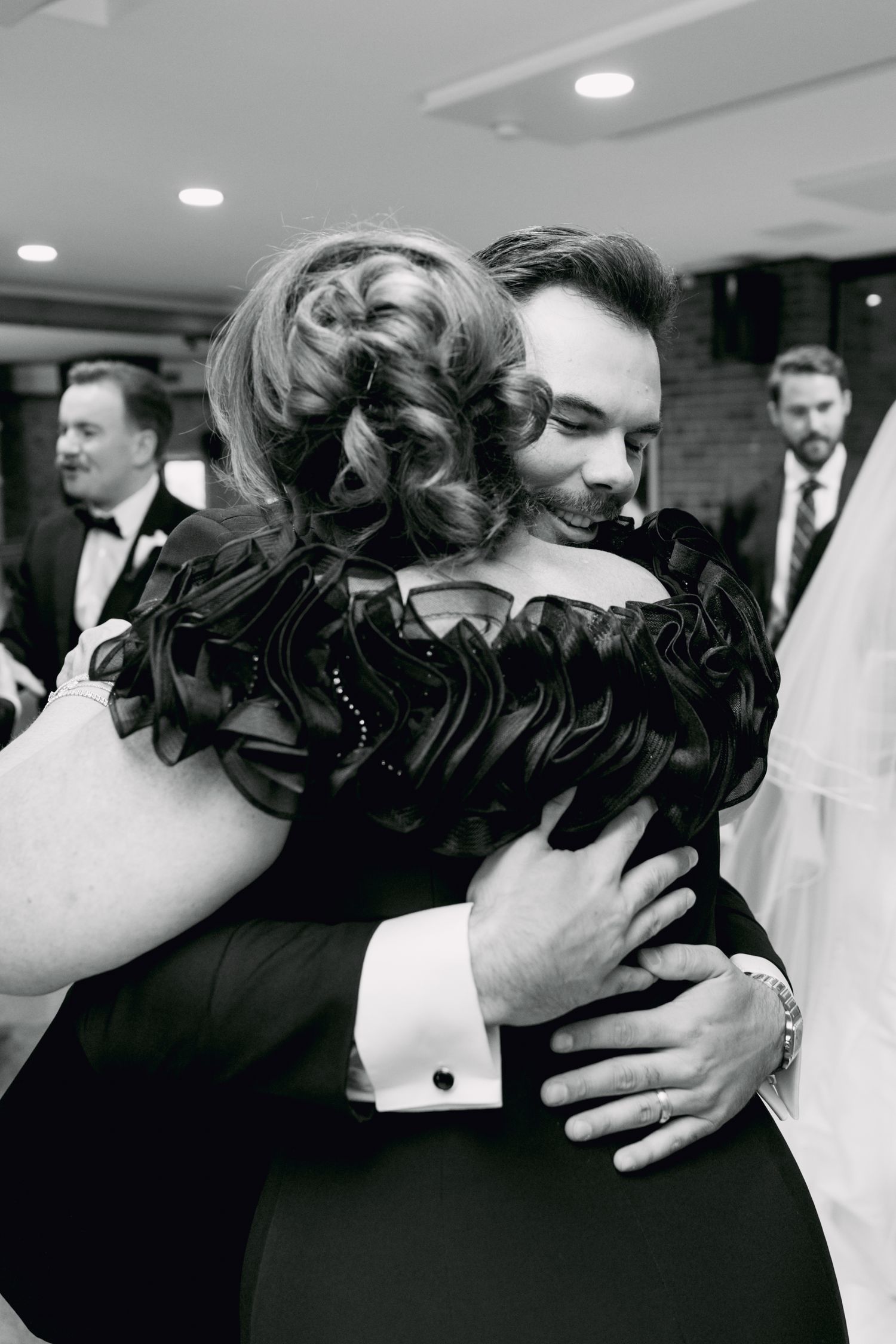 Black and white photo of a groom hugging a woman in a black dress at a wedding reception.