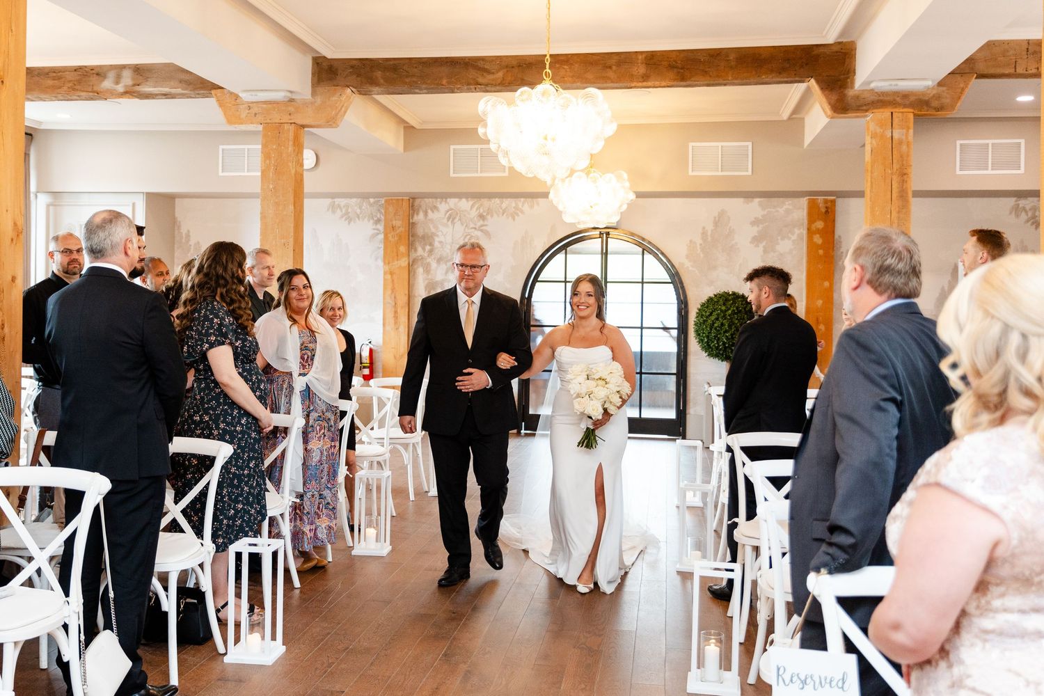 Bride walks down the aisle with her father in an elegant indoor wedding venue with white chairs and chandelier.