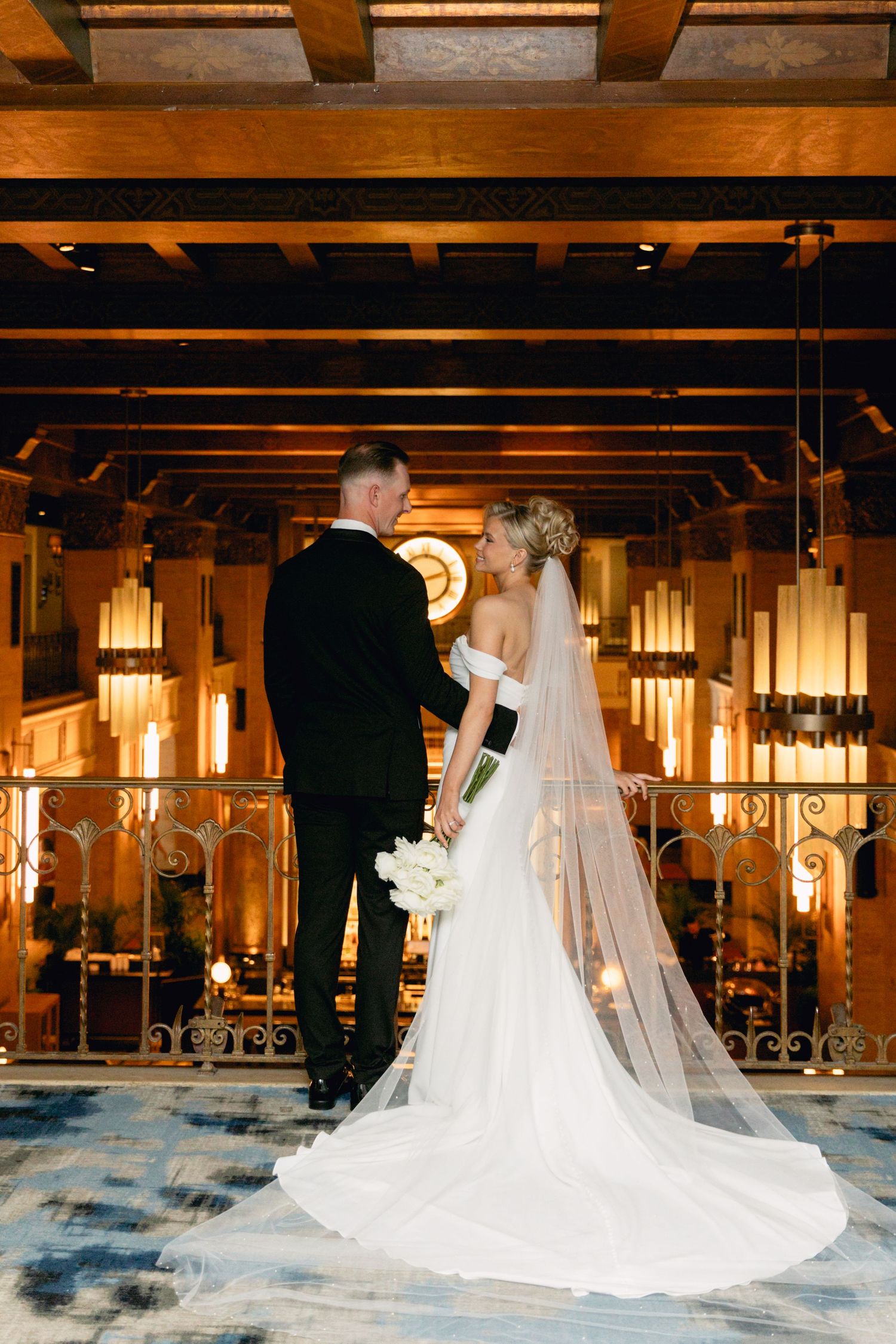 Bride and groom stand together on elegant balcony, her lace veil trailing on patterned floor in warmly lit Art Deco venue.