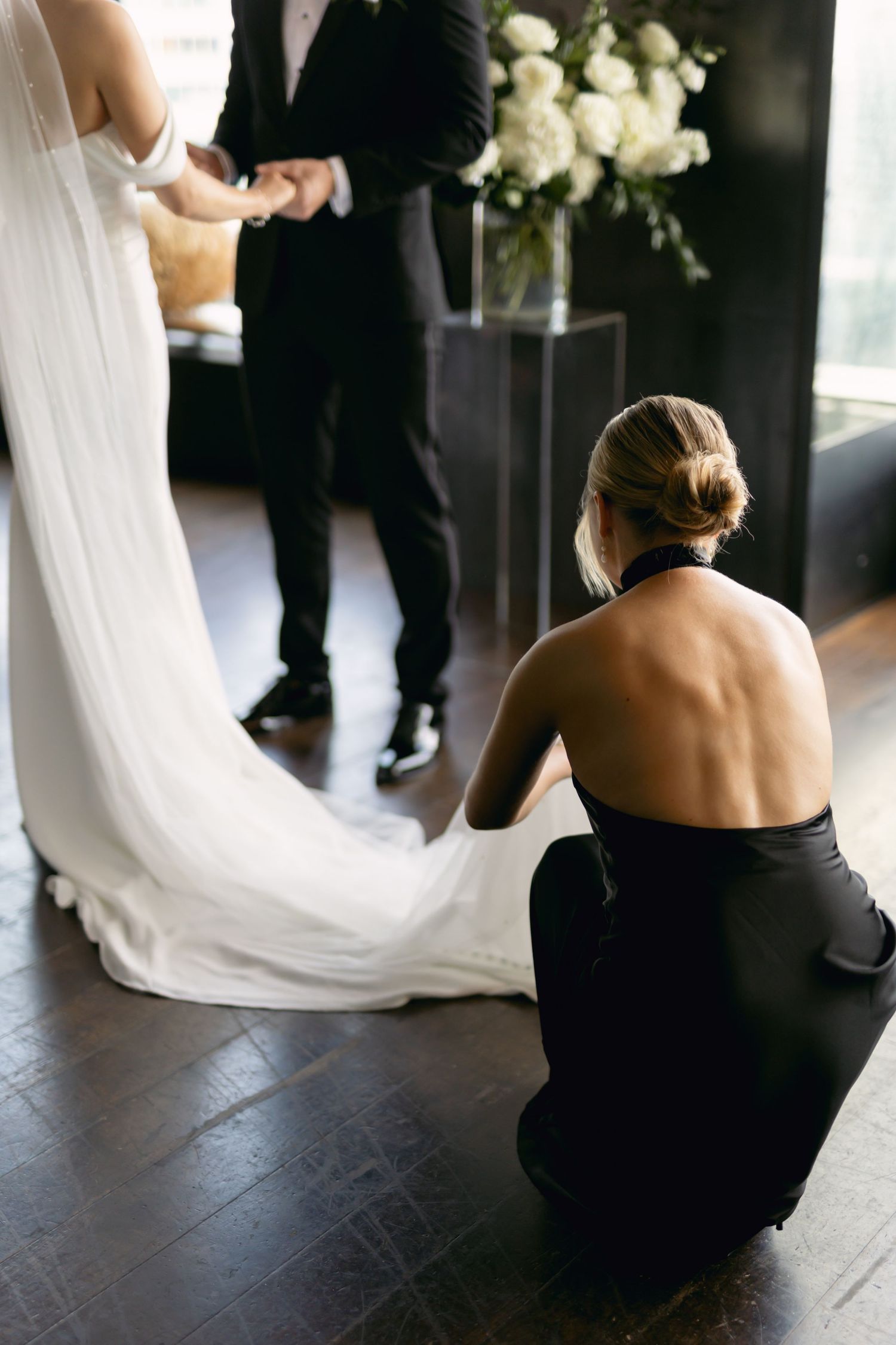 Bridesmaid in black backless dress kneeling to arrange bride's white wedding gown train before ceremony.
