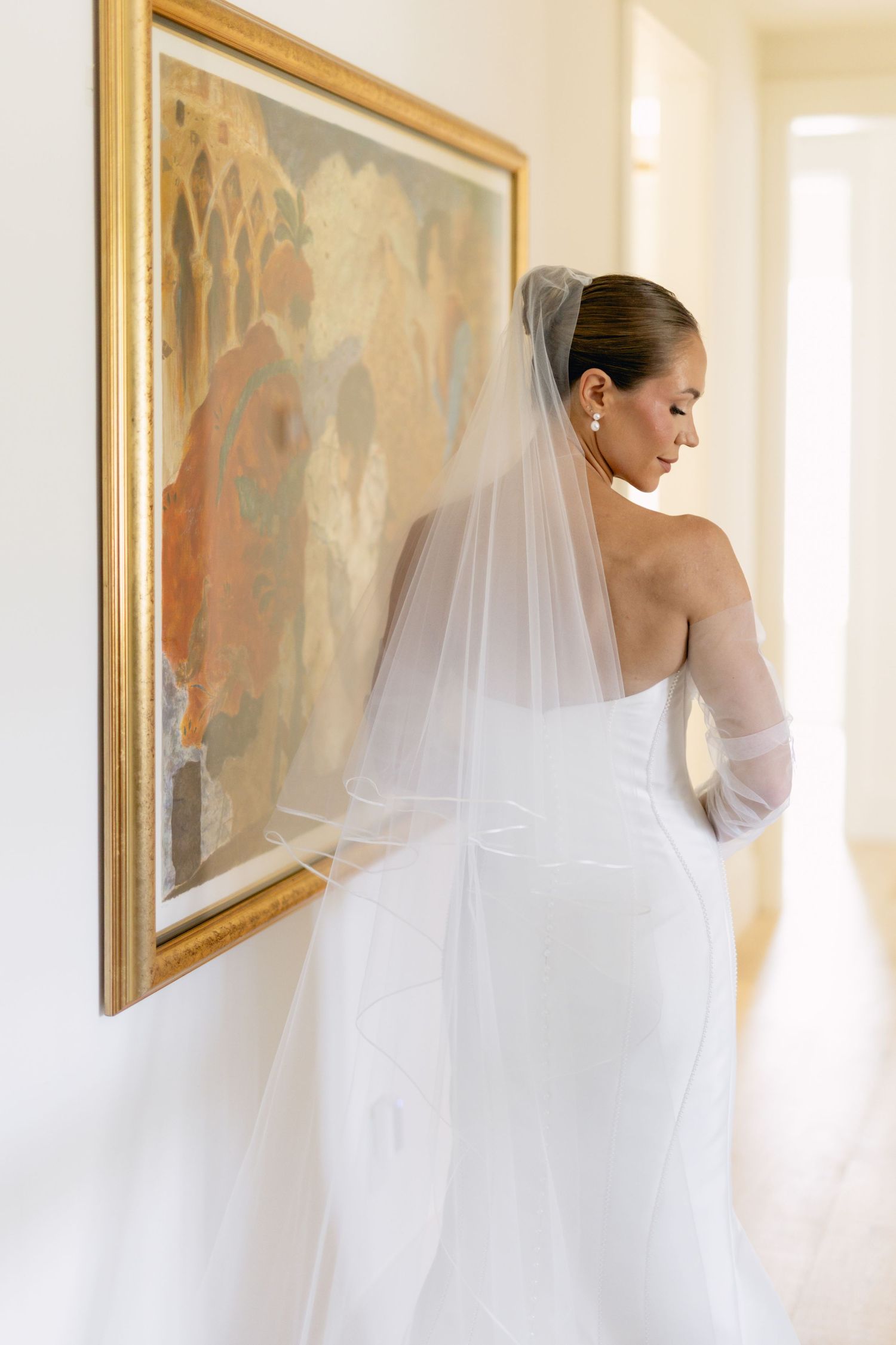 Elegant bride in strapless white gown with long veil standing beside gold-framed artwork indoors.