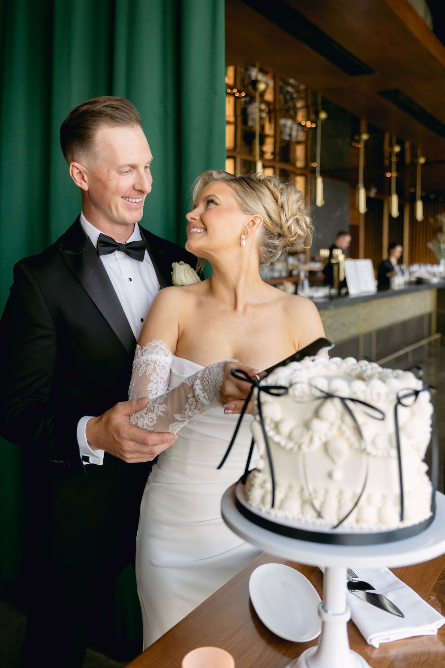 Bride and groom smile together preparing to cut elegant white wedding cake with black bow detail.