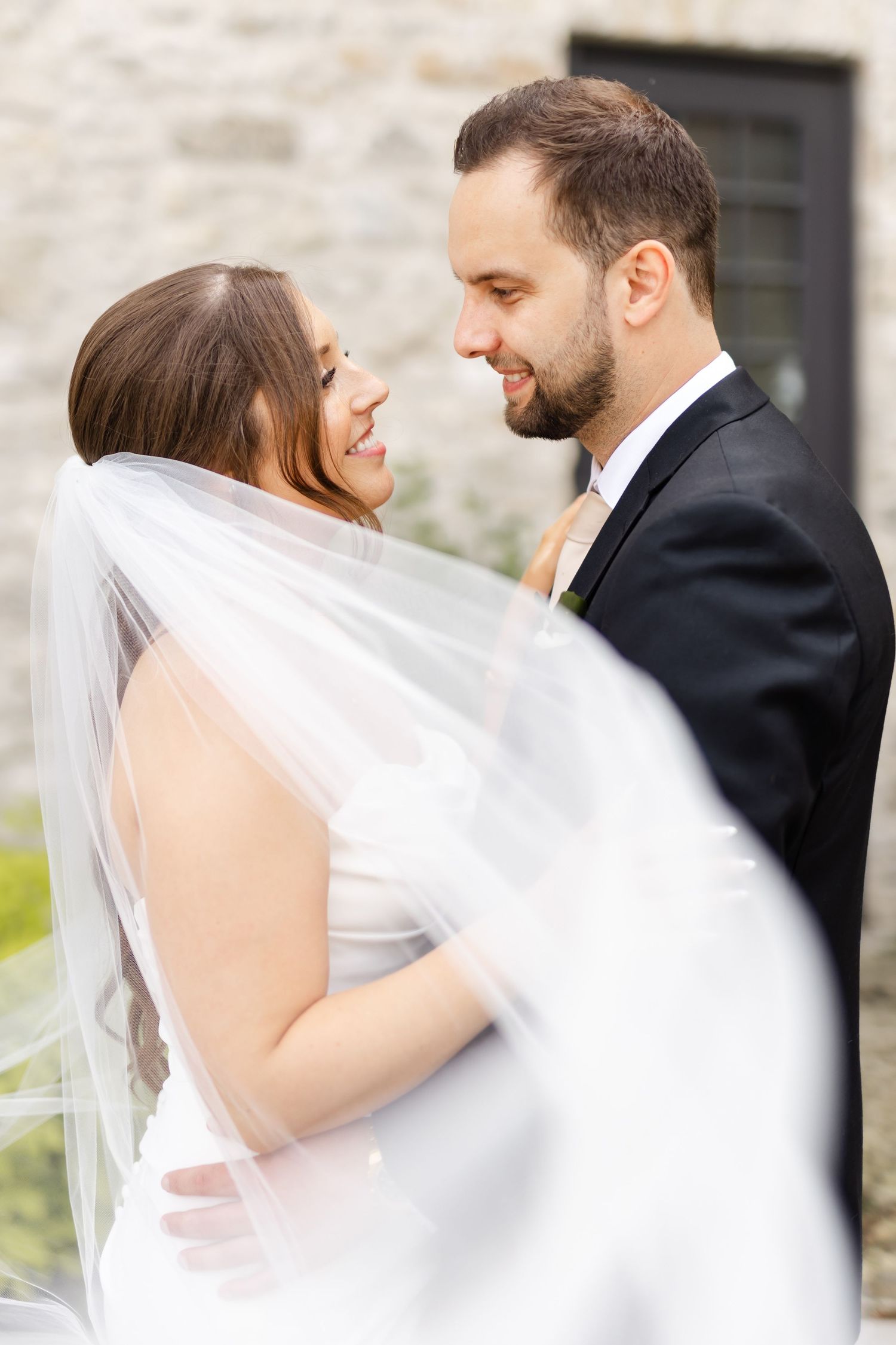 Bride and groom share an intimate moment smiling at each other, bride's veil flowing in the foreground.