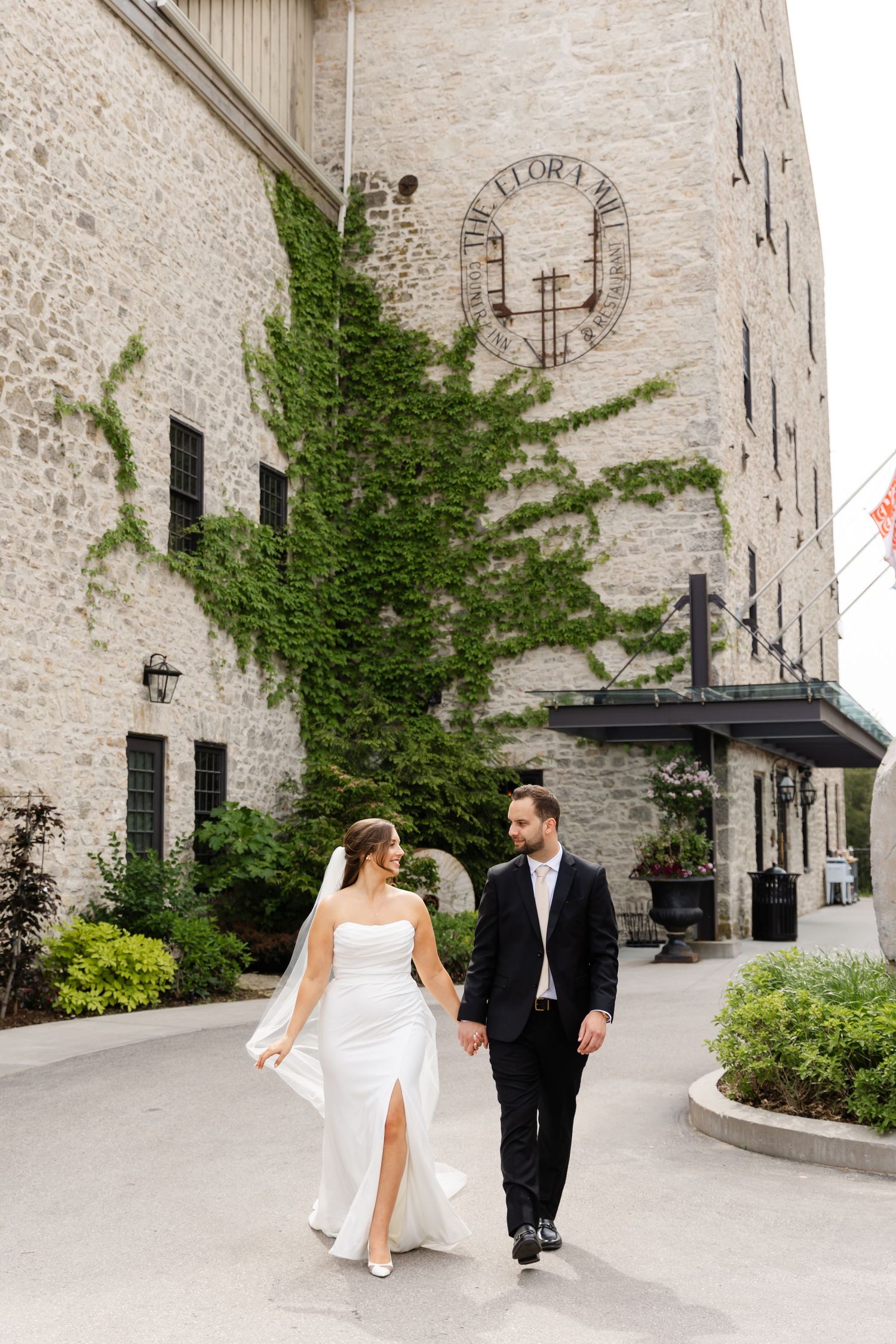 Bride and groom walk hand in hand outside historic stone building with ivy-covered walls.