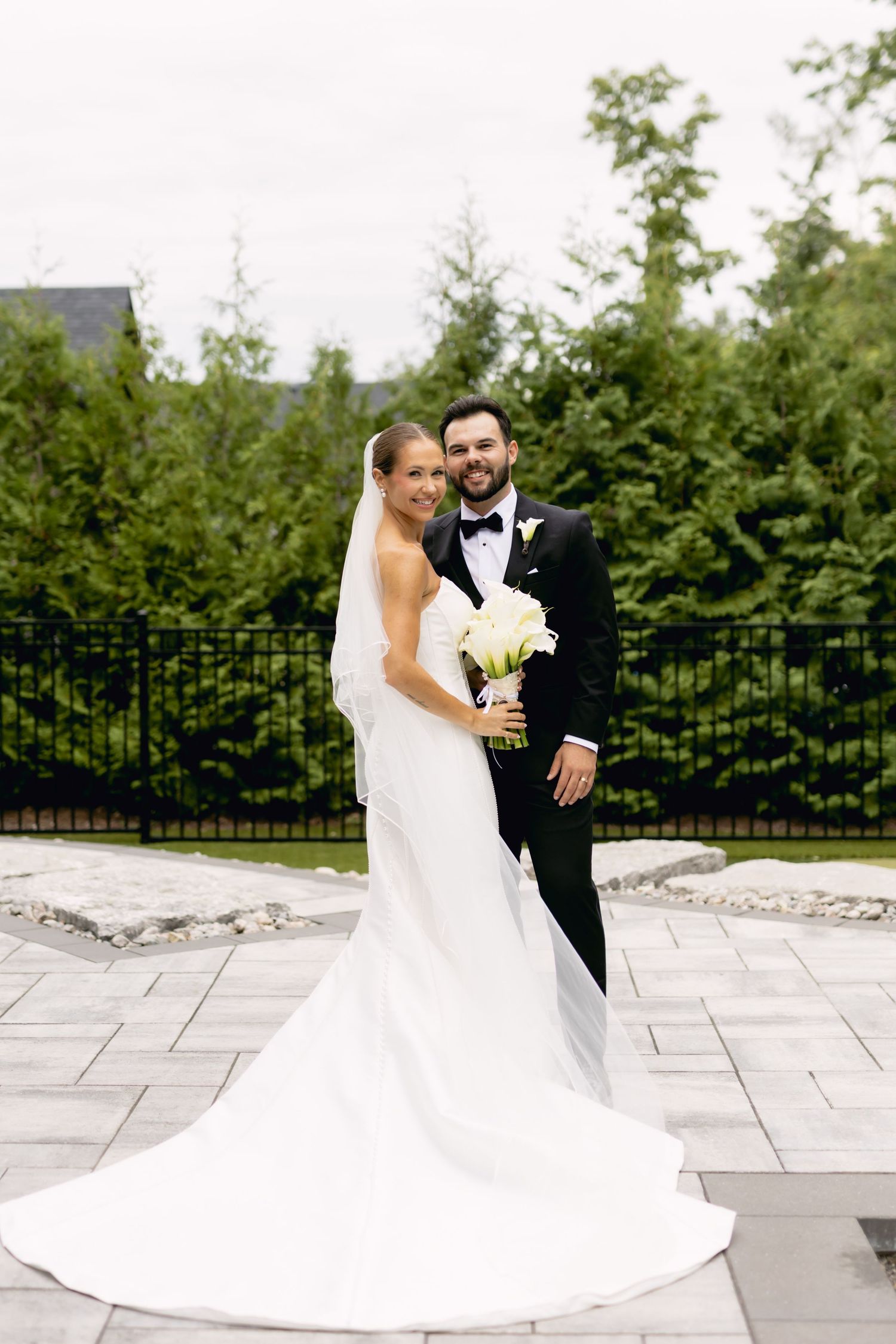 Bride and groom pose together outdoors, she in white gown with train, he in black tuxedo.