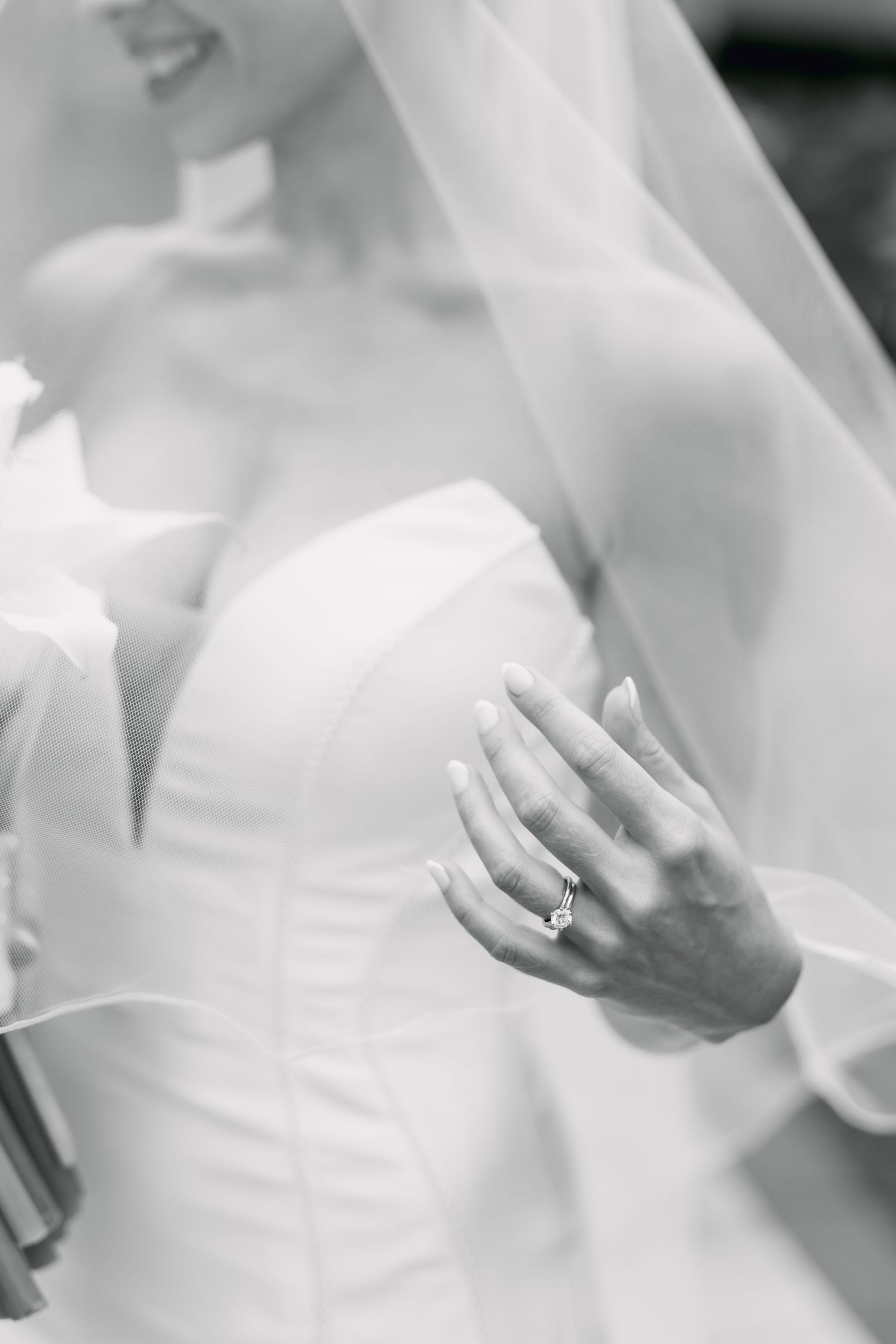 Black and white close-up of bride in strapless gown displaying diamond engagement ring under veil.