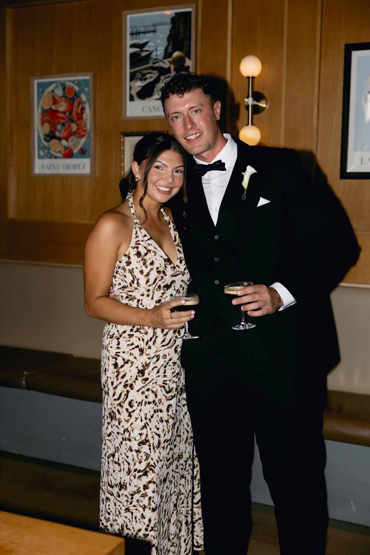 Couple in formal attire holding champagne glasses pose together at a wedding reception venue.