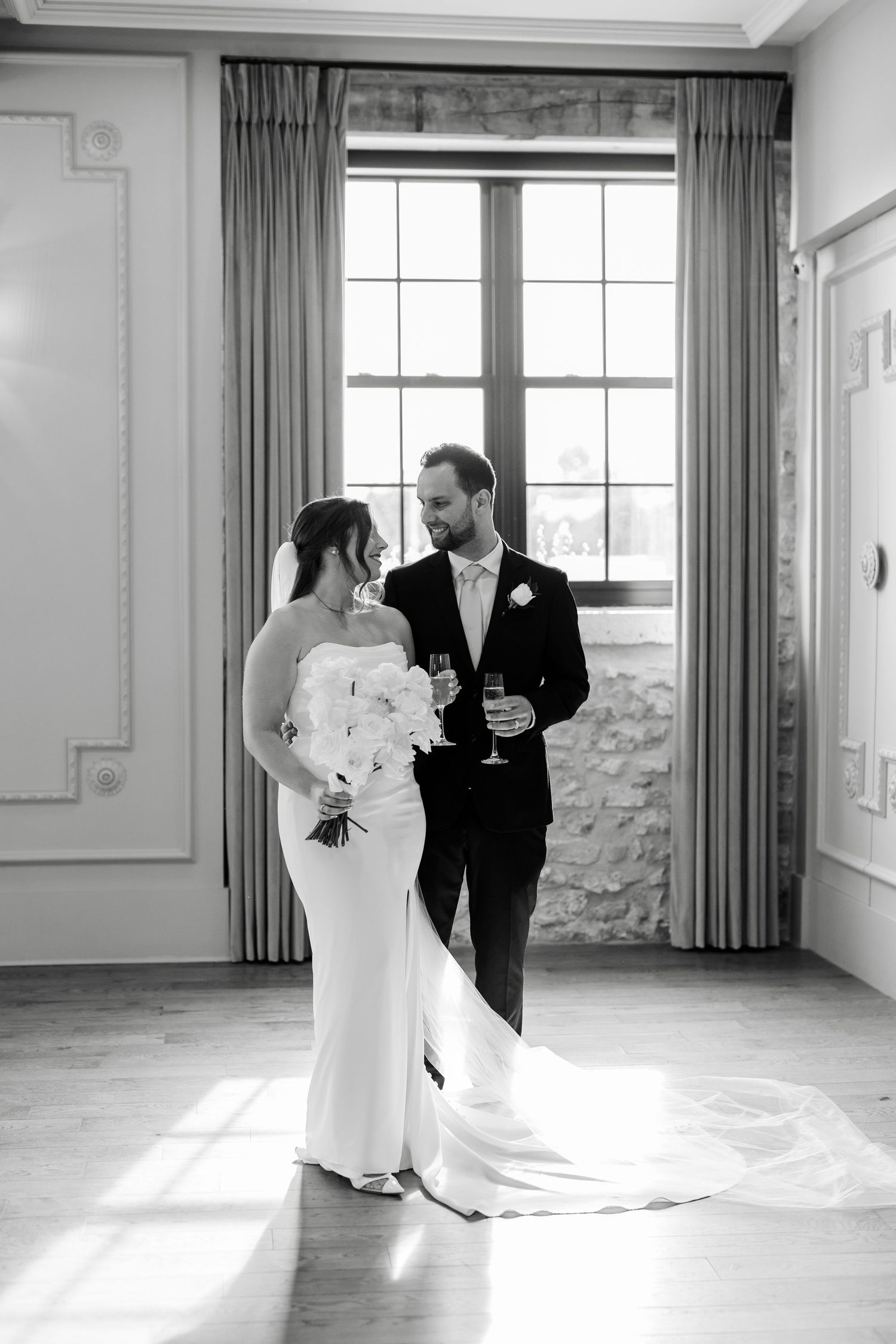 Black and white portrait of bride and groom holding champagne glasses by a sunlit window in elegant venue.