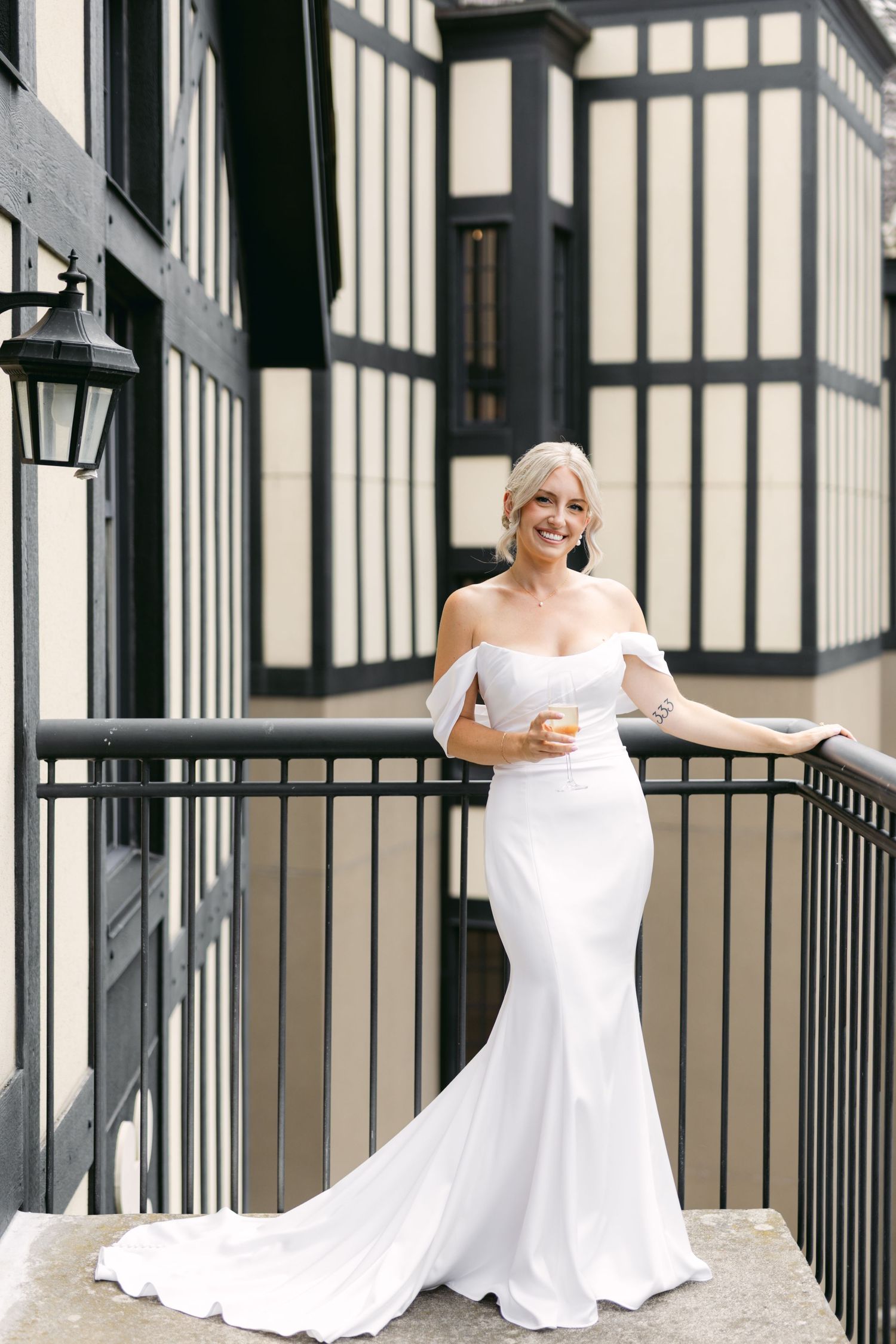 Bride in a white off-shoulder wedding dress smiling while sitting on a balcony with black railings.