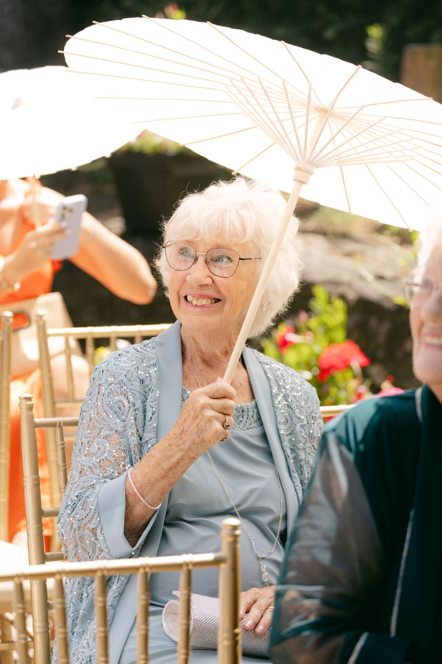 A wedding guest holding a white parasol while seated in a garden setting.
