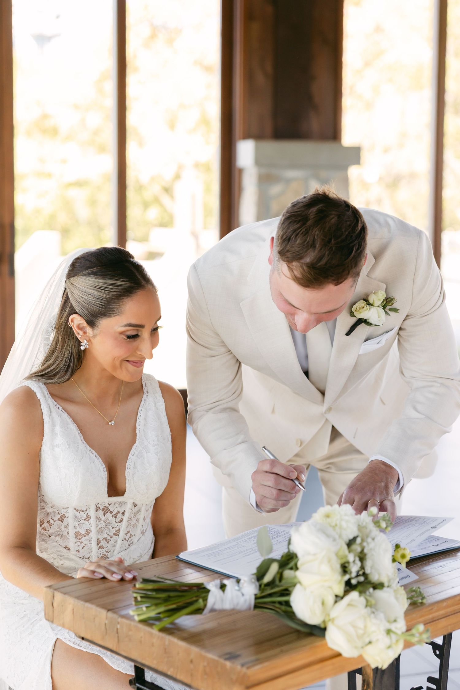 Couple signing marriage documents on a wooden table during wedding ceremony.