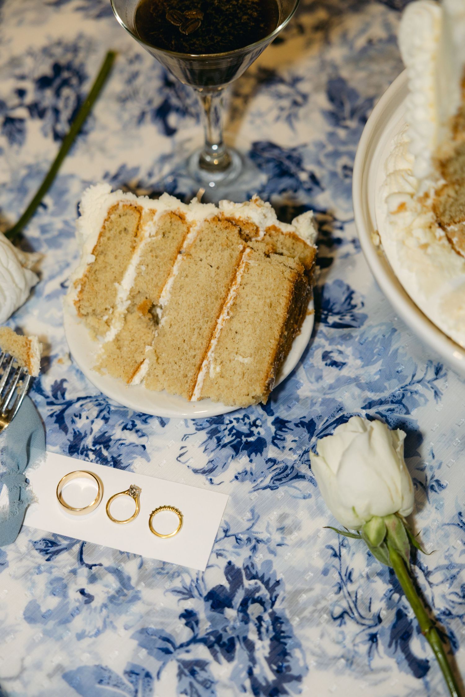 Slice of layered vanilla wedding cake with wedding rings and white roses on blue floral tablecloth.
