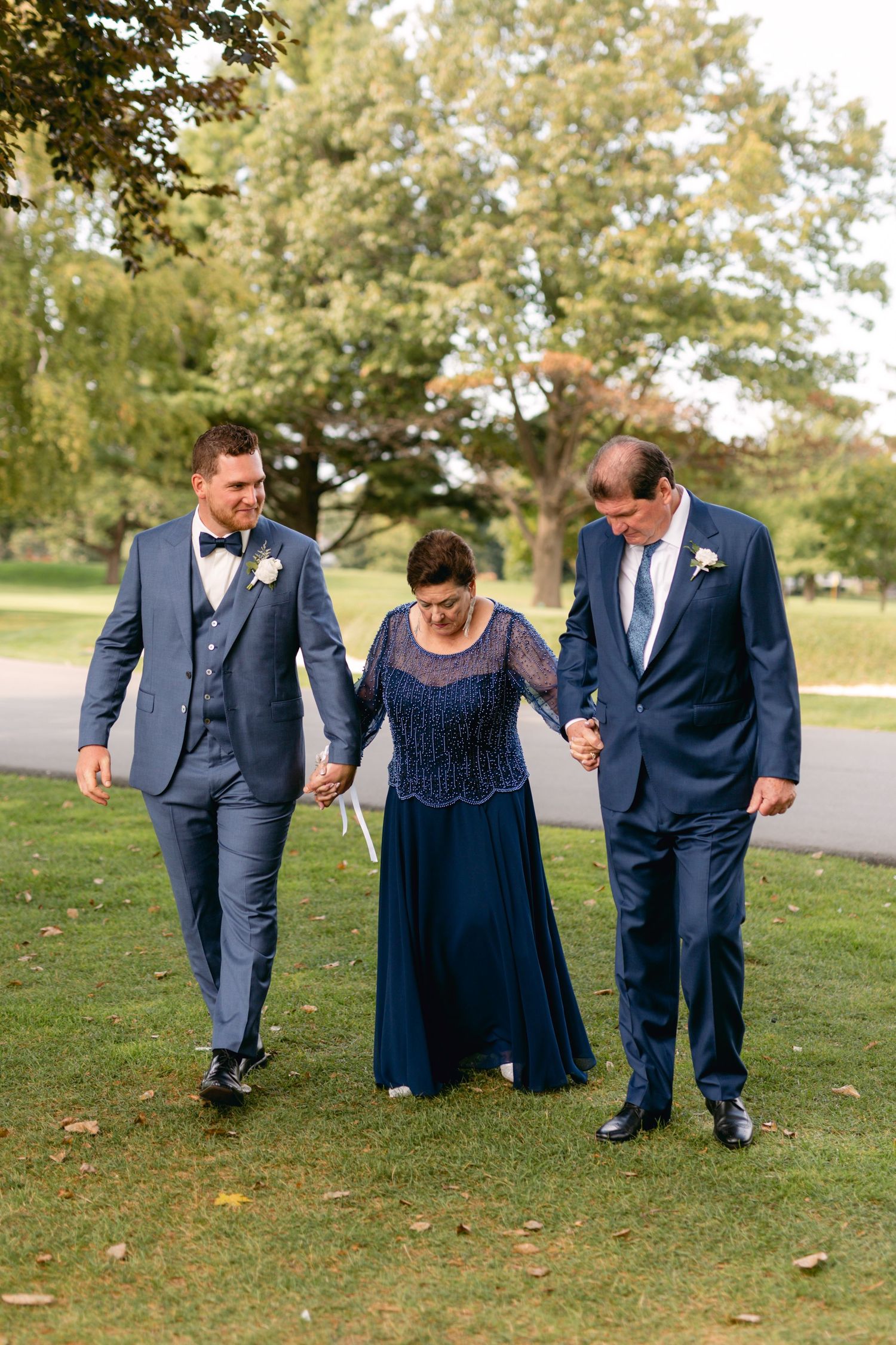 Three people walking arm in arm on grass during wedding celebration.