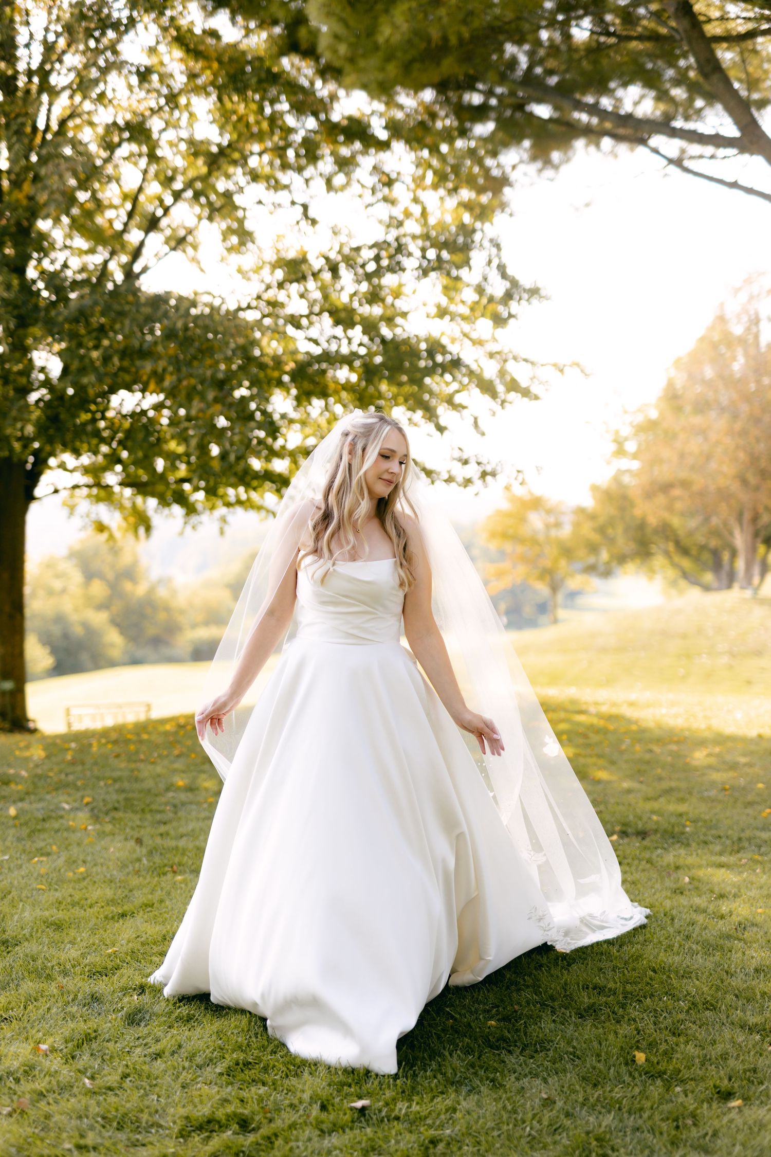 Bride in flowing white dress twirls on grassy hillside during sunset.